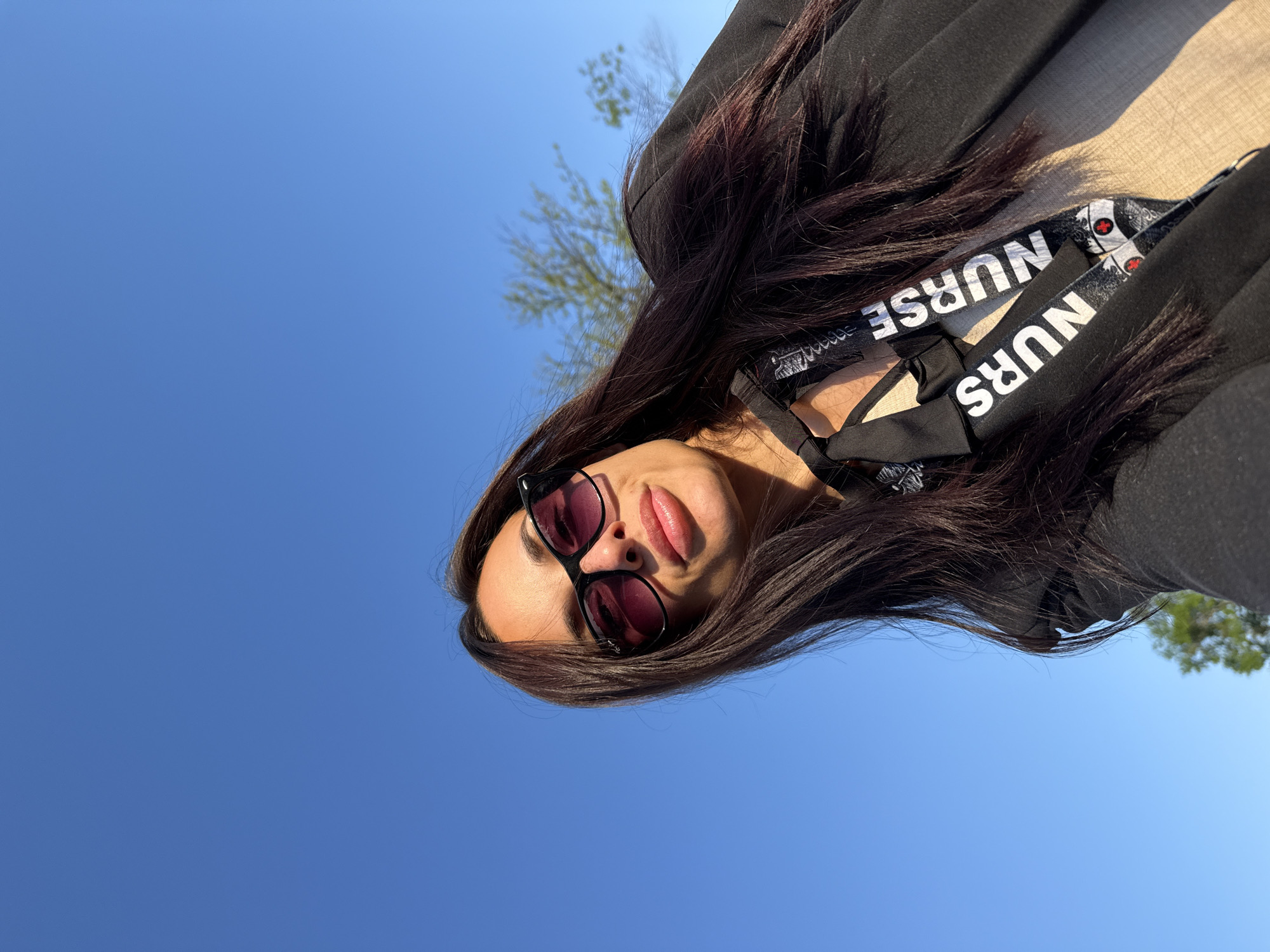 A woman is shown in a tilted, upward-facing shot against a clear blue sky. She is wearing sunglasses and a lanyard with the word "NURSE" printed on it. She has long, dark hair. The background shows hints of trees. The location of the city of Pleven, Bulgaria cannot be determined from the image.