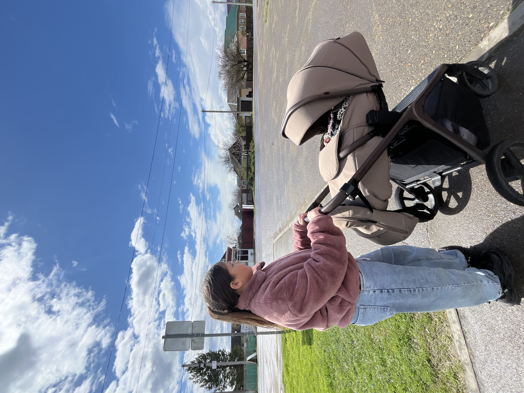 A woman in a pink jacket and blue jeans is standing on a paved road, pushing a beige stroller. The location is identified as Novopokrovskaya, Russia, based on visual clues such as architectural style and signage. The woman is looking ahead, presumably at the path in front of her. The stroller is positioned in front of her, and the scene includes a clear blue sky with scattered clouds, houses, and a street sign. The woman's black shoes are visible as well. Patches of grass are visible on one side of the road.