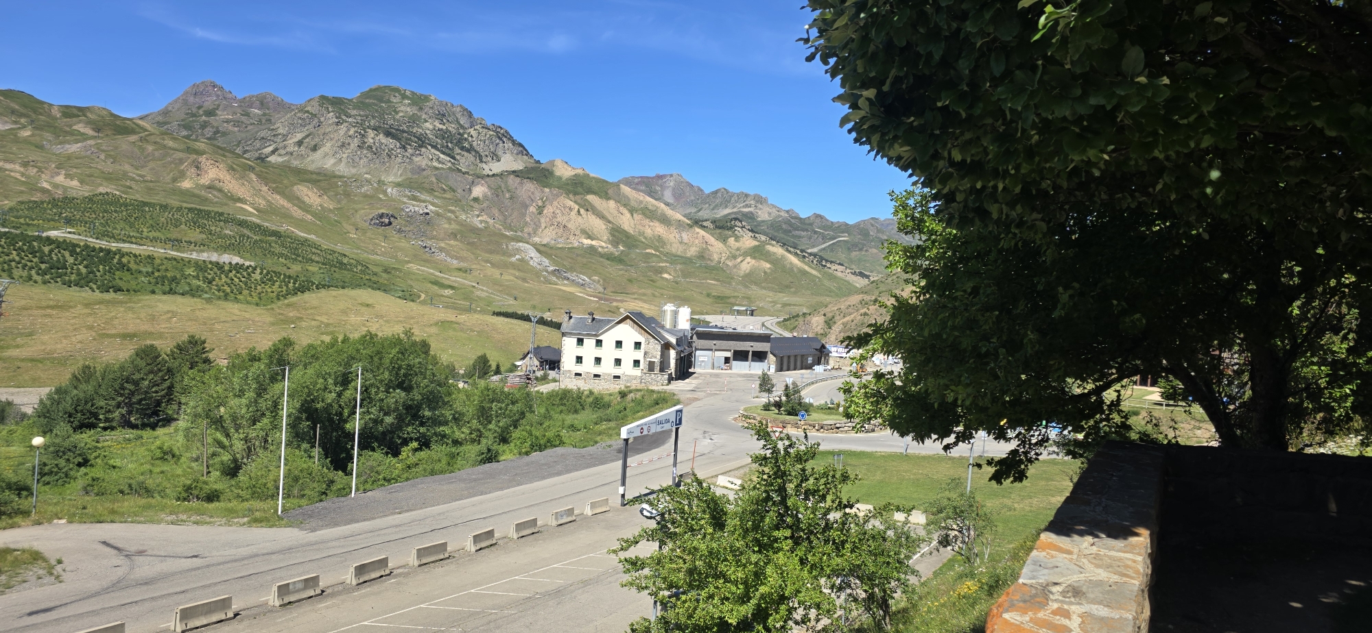 The image depicts a mountain landscape with a road and buildings in the foreground. A bright blue sky and sunny weather suggest it is daytime. The scene is set in a mountainous region, possibly a ski resort or a mountain village, given the presence of a road leading towards buildings and the expansive, undeveloped hillsides.
In the middle ground, several buildings are visible. One prominent building is a light-colored, multi-story structure with a dark roof, featuring several windows. Adjacent to it are other low-rise buildings, some with large, open entrances, suggesting they might be service or storage areas. A paved road curves through the area, with white concrete barriers lining its edge. There are also signs visible on the side of the road, including a sign with the word "SALIDA" (exit in Spanish) and a parking symbol.
The foreground is partially obscured by the branches and leaves of a large green tree on the right side of the image. The hills in the background are covered in green grass and dotted with trees, with some areas showing bare rock or patches of lighter-colored earth. The terrain appears steep and rugged, characteristic of mountain environments. In the far distance, more mountains are visible, their peaks reaching towards the sky. The overall impression is one of a serene and vast natural landscape with man-made structures integrated into it. The time of day appears to be late morning or early afternoon due to the strong sunlight and clear shadows.