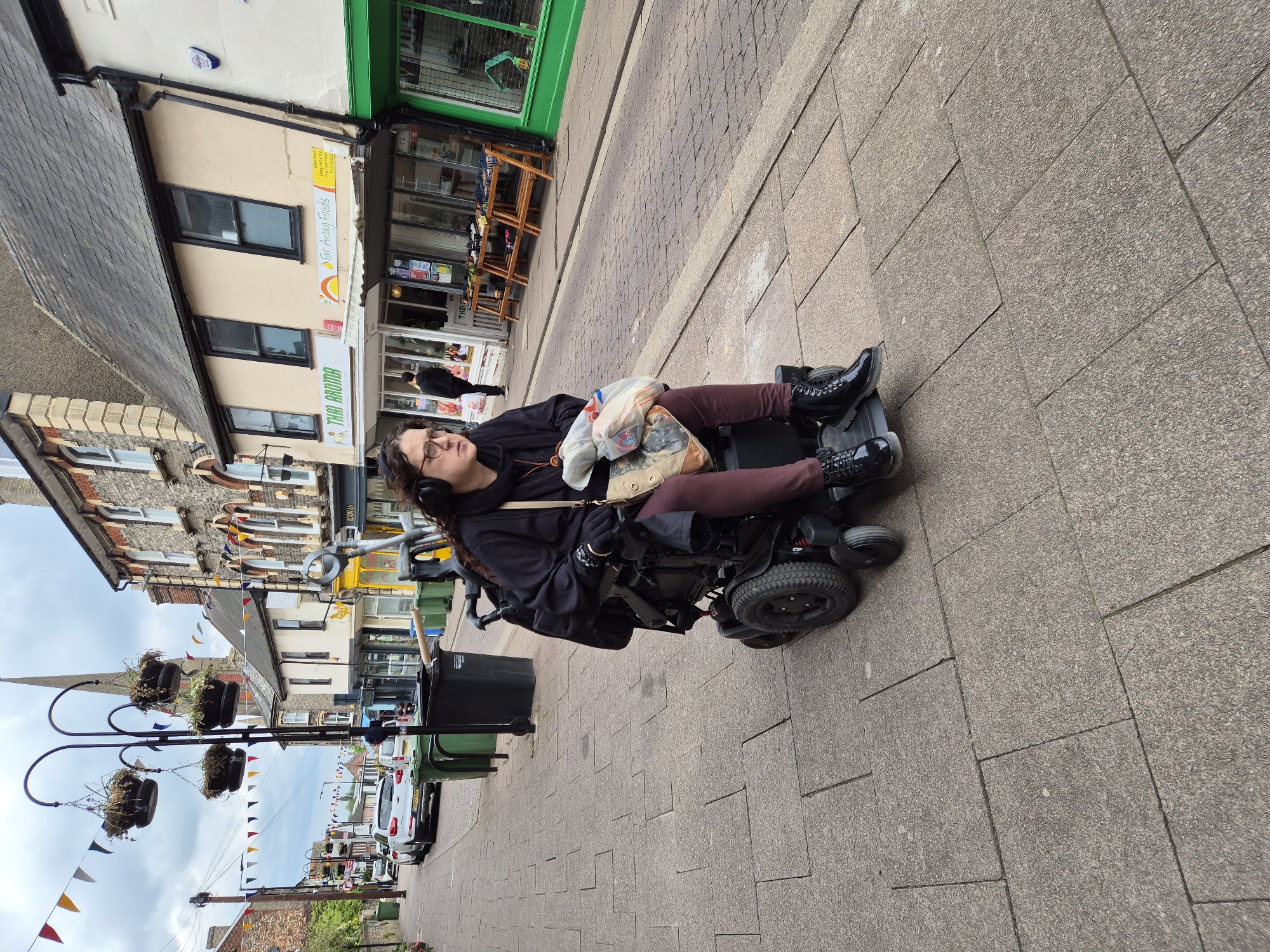 A woman with curly dark hair and eyeglasses is seated in a powered wheelchair on a sidewalk in a street scene. She wears a black hooded top, maroon pants, and black boots. Bags are placed on her lap. The sidewalk is paved with rectangular gray stones. Behind her is a street with buildings, some adorned with bunting, suggesting a celebratory or festive atmosphere. There are parked cars on the left side of the road. The location is in the city of West Suffolk, United Kingdom.