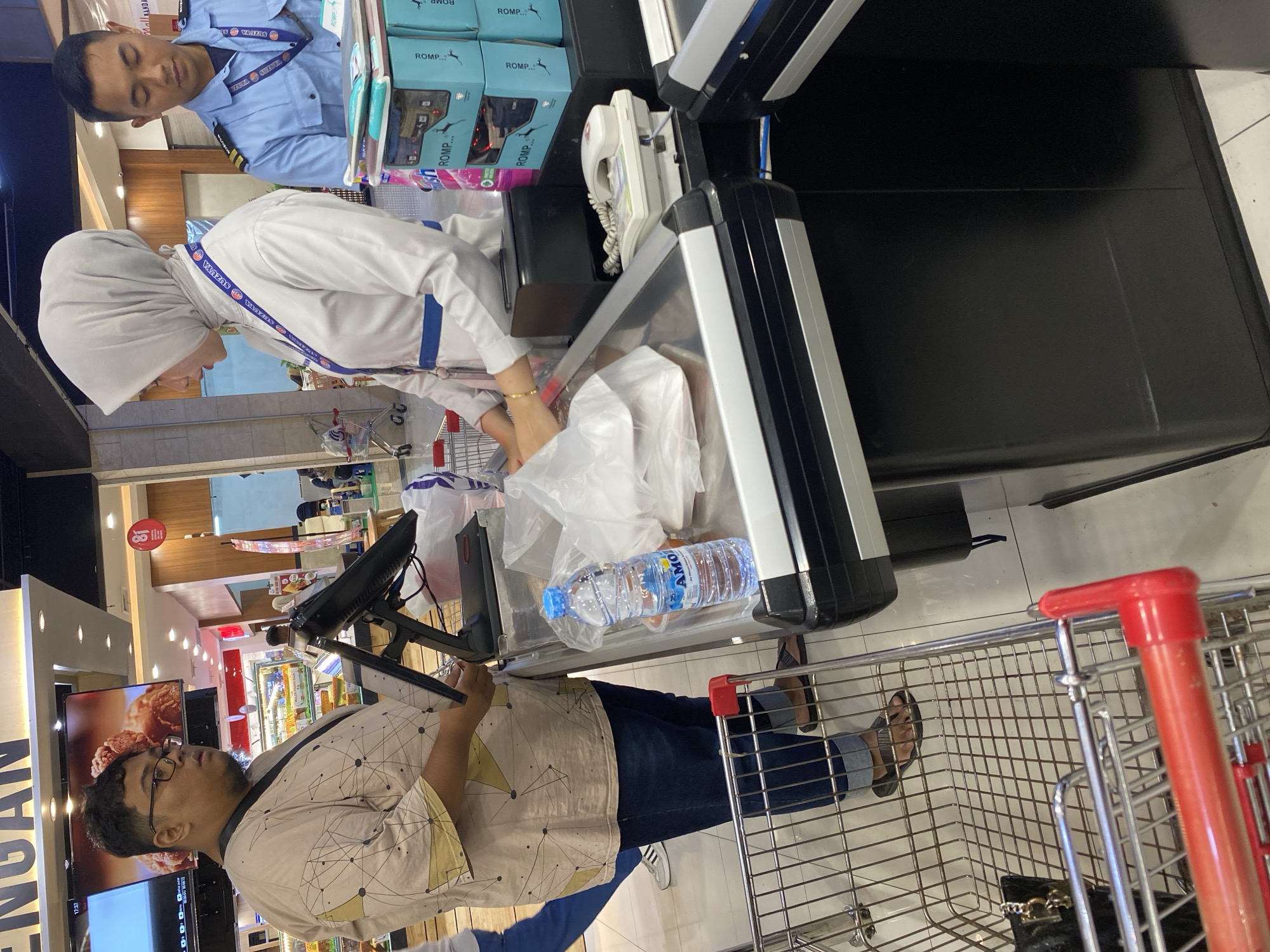 The image captures a scene at a checkout counter in a retail environment, likely a supermarket, located in Banda Aceh, Indonesia. A person, identified by their attire as a security guard or staff member, is visible in the background on the right, wearing a light blue uniform with epaulets and a dark blue tie. In the foreground, a customer, a man wearing glasses and a patterned beige shirt over a dark t-shirt and blue jeans, is standing at the counter. He appears to be holding plastic bags filled with purchased items. To the left of the customer, a red-handled shopping cart is partially visible, containing a black bag. A cashier, a woman wearing a light gray hijab and a white uniform with a blue sash, is attending to the customer. She is in the process of scanning or bagging items. On the counter, several objects are present, including a black monitor, a white landline telephone, a bottle of "AMO" brand drinking water, and stacks of boxes with "ROMP" branding. The environment suggests an indoor commercial space, with lighting and promotional displays visible in the background.