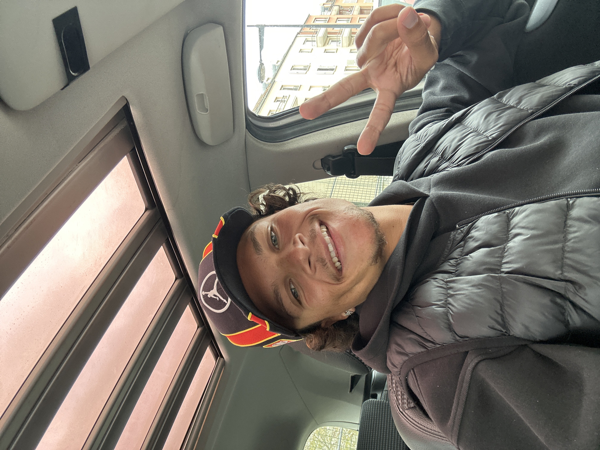 A man is taking a selfie inside a car. He is wearing a black hoodie, a black puffer jacket, and a baseball cap with a black, red, and gold design, featuring a logo that includes a stylized figure. He is smiling and making a hand gesture. The car has a sunroof with a louvered sunshade. Outside the car window, a multi-story building is visible. The scene is located in Noisy-le-Grand, France.