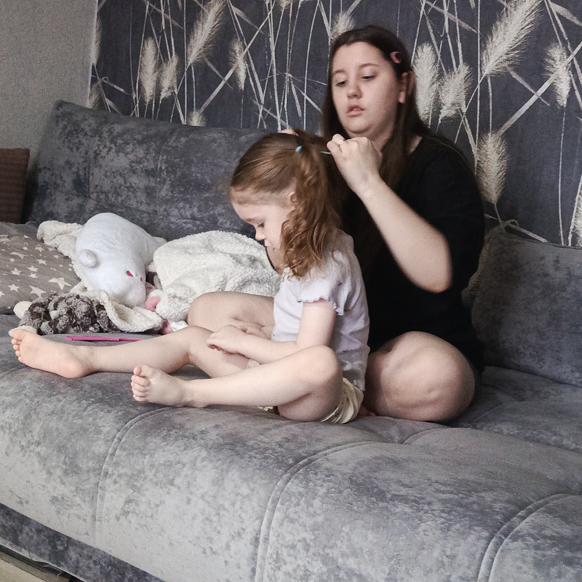 A young woman with long brown hair is braiding the hair of a young girl with reddish-brown hair, in Марковский, Russia. The girl sits cross-legged on a grey velvet couch with a throw pillow and stuffed animals. The woman sits behind the girl and is actively styling the girl's hair. The backdrop is a wall with a patterned wallpaper.