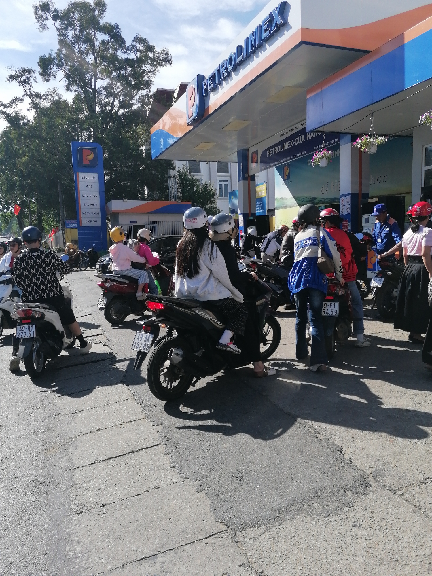 The image depicts a busy scene at a Petrolimex gas station in Da Lat, Vietnam. Several people on motorcycles are waiting in line, some possibly to refuel or make a purchase. In the foreground, a person wearing a black and white patterned shirt and black shorts is seated on a white motorcycle, facing away from the camera. To their left, another white motorcycle is visible. In the center, a person in a white jacket and black helmet, and another person in a blue and white jacket and blue helmet are on a red motorcycle. To the right, a woman in a pink shirt and black skirt stands near a motorcycle, and a man in a blue uniform, likely an employee of the gas station, is also present. The gas station features prominent "PETROLIMEX" signage and a tall blue and white sign listing services like "XĂNG DẦU" (gasoline), "GAS", "DẦU NHỚN" (lubricant), "BẢO HIỂM" (insurance), and "DỊCH VỤ" (service). The environment suggests a warm, sunny day with clear skies and scattered clouds, casting shadows from the people and motorcycles onto the cracked asphalt ground. Trees are visible in the background. The overall atmosphere is one of everyday commerce and activity.
