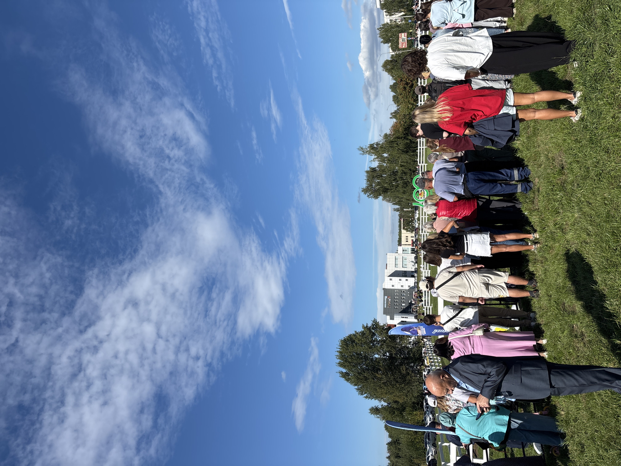 This image depicts an outdoor gathering of people on a grassy field under a partly cloudy blue sky. A modern, multi-story building with a grey facade and numerous windows is visible in the background, surrounded by trees. A white fence runs horizontally across the middle ground. Several people are standing and facing away from the camera, indicating they are looking at something or someone beyond the frame. Some individuals are wearing casual clothing, while others are dressed more formally. Blue flags with white designs are planted in the ground. A large green sign with white lettering is also present in the background, though the text is partially obscured. Cars are parked in the distance behind the fence. The overall impression is of a public event or festival taking place on the outskirts of a city. The specific location is likely a park or event space in Naberezhnye Chelny, Russia, given the architectural style of the building and the presence of Russian text on some of the flags.