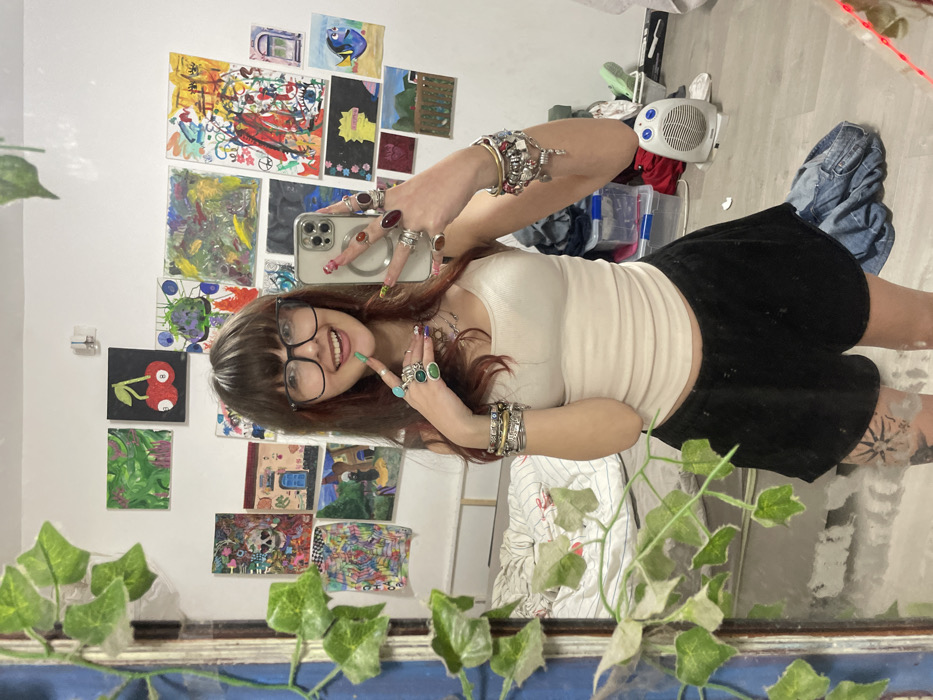 A smiling person with brown hair and bangs, wearing glasses, a beige top, and black shorts, poses for a selfie in a mirror. Multiple rings and bracelets adorn their hands. Behind them, a wall is decorated with various artworks. A white heater sits amidst clutter, including boxes and clothes. Artificial vines frame the bottom of the mirror. A tattoo is visible on the person's leg.