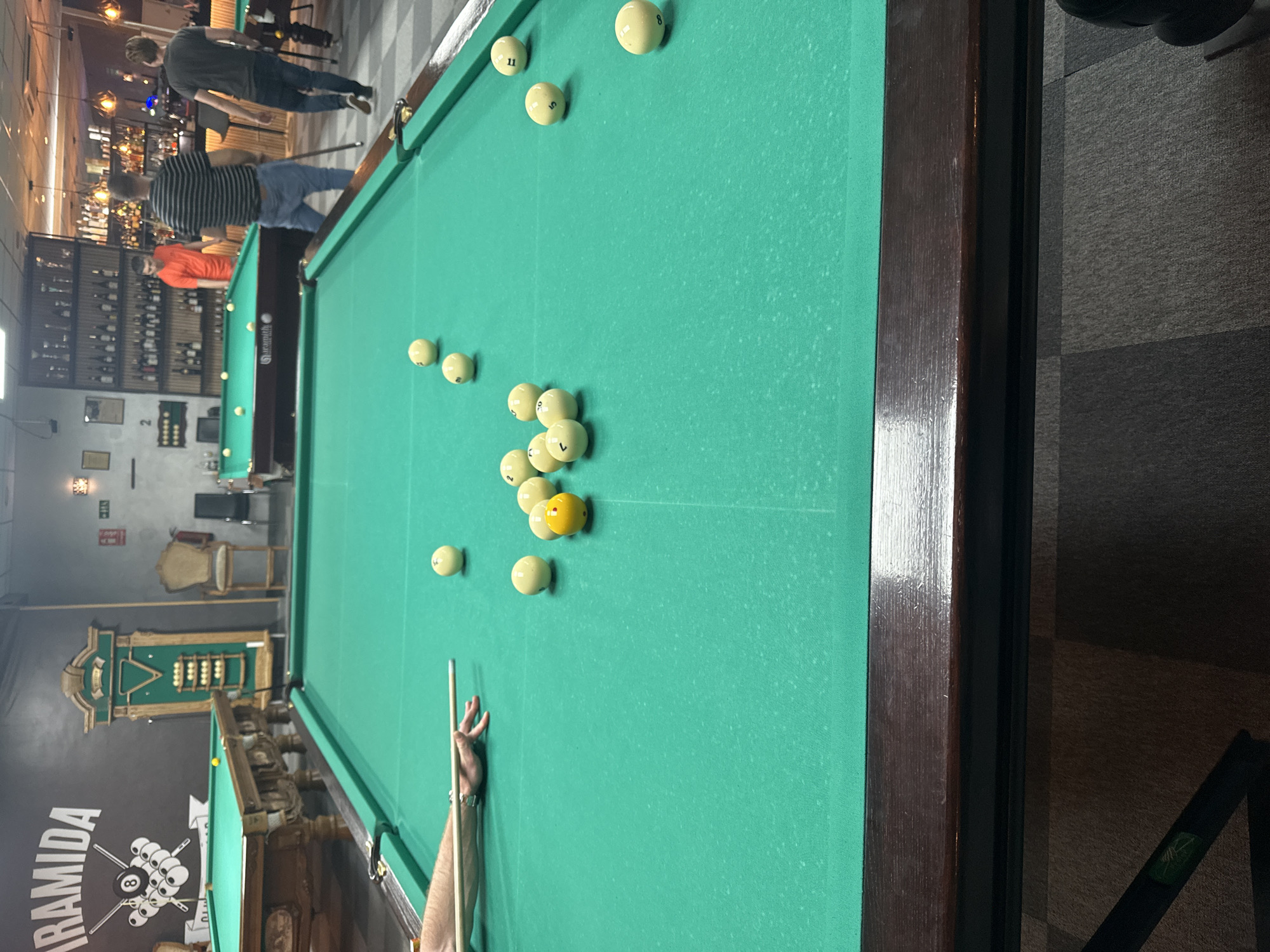 The image depicts a billiards game in progress in a bar or game room. A pool table dominates the scene, covered in green felt and scattered with multiple white balls and one yellow ball. A person's hand rests on the table next to a cue stick, suggesting an imminent shot.
In the background, other people are present, moving around in the bar area. A well-stocked bar is visible, featuring shelves filled with various bottles. The branding "Piramida" is displayed on a wall near another pool table.
There is no specific evidence to confirm the location as San Vicente Del Raspeig/Sant Vicent Del Raspeig, Spain, and therefore the location cannot be verified with certainty.