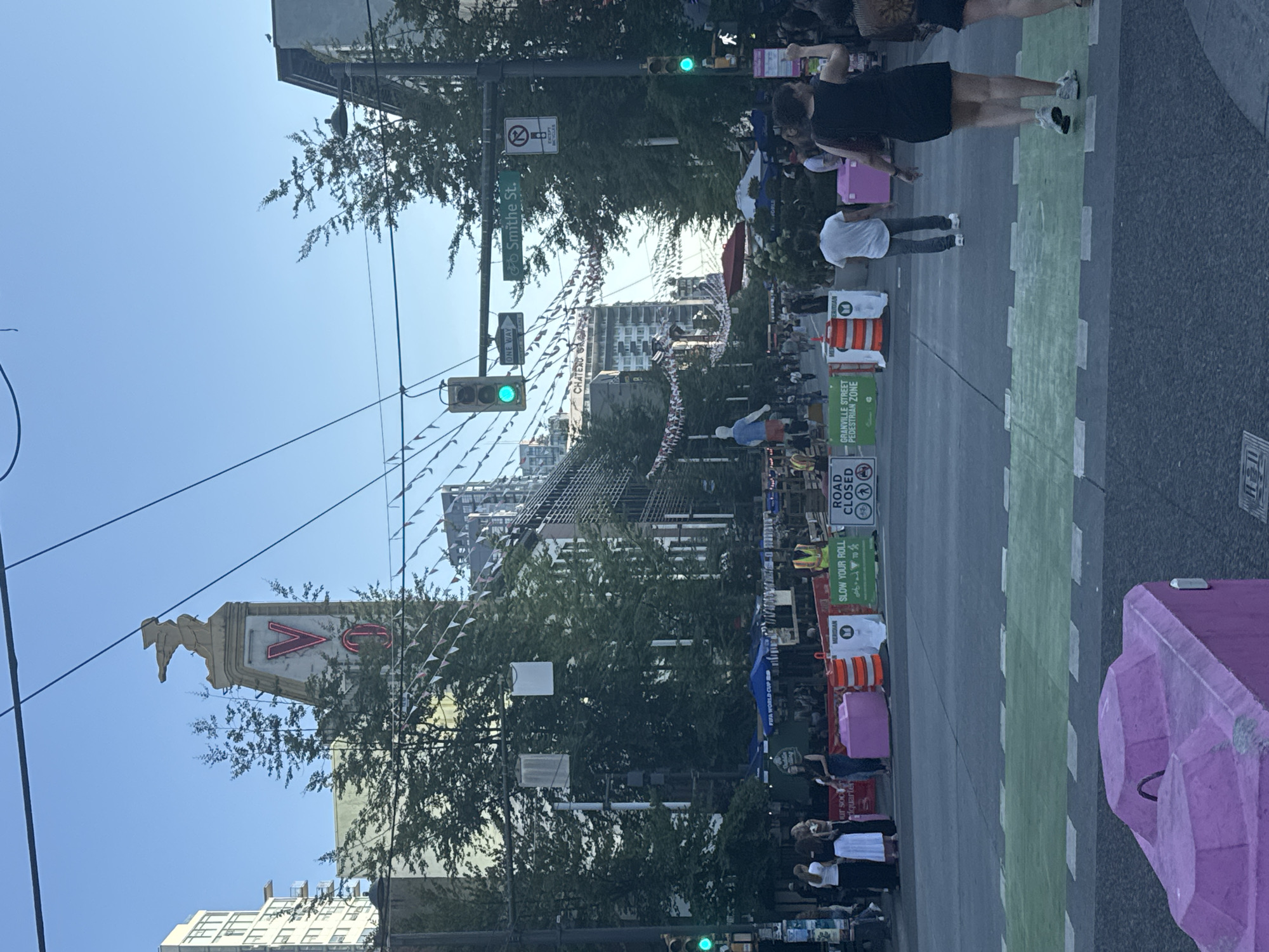 The image captures a street scene in Vancouver, Canada, likely during an outdoor event or festival. In the background, a prominent neon sign with a stylized "V" is visible atop a building, possibly identifying a landmark like the Vogue Theatre, which is known for its historic signage and location in Vancouver's downtown core. The street is decorated with triangular flags strung across it, suggesting a festive atmosphere. Traffic lights and street signs, including one indicating "Smithe St.", are present, as are various trees and modern buildings, typical of an urban environment.
Several people are present in the image, some walking and others standing in groups. Orange and white traffic barriers and larger pink concrete barriers are set up, indicating a street closure or a designated pedestrian zone. Signs visible include "ROAD CLOSED," "GRANVILLE STREET PEDESTRIAN ZONE," and "SLOW YOUR ROLL." In the foreground, a green-painted crosswalk is a dominant feature, with a large pink concrete barrier in the immediate foreground. The overall impression is of a lively urban public space adapted for pedestrian use and a special occasion.