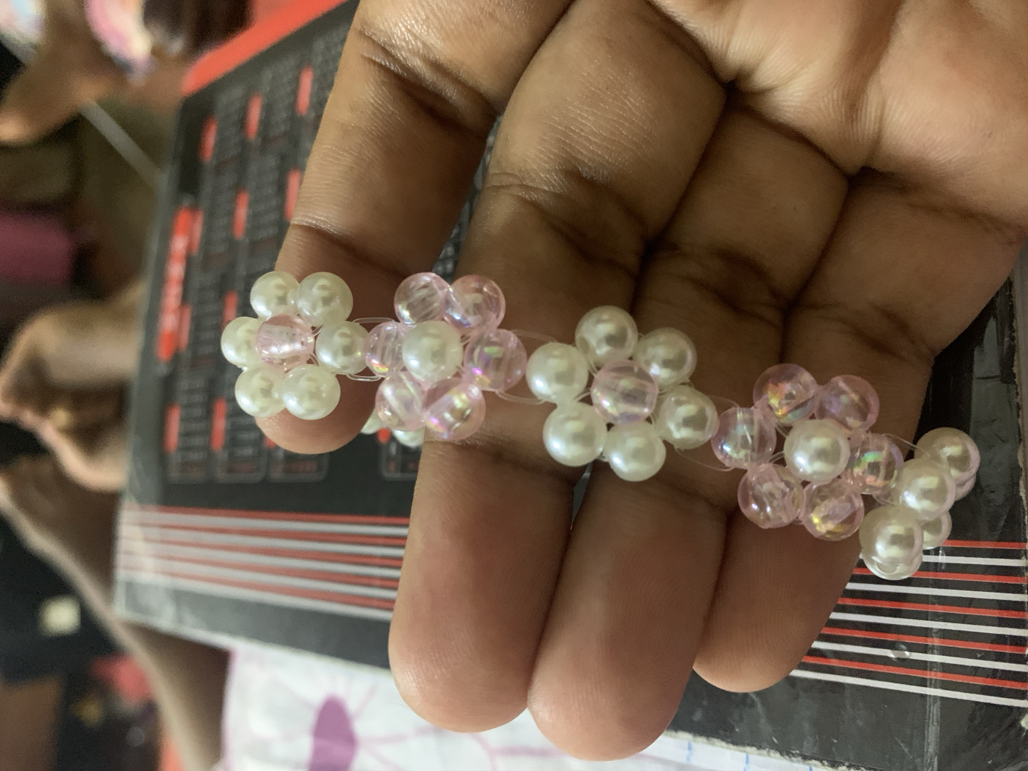 A hand, with brown skin, is holding a beaded ring/bracelet. The ring/bracelet is made up of small beaded flowers that alternate in color; white and light pink. The background is blurry. There is a black object with red and white stripes. There is also a white piece of tissue paper with a purple stain. The location cannot be determined based on the image.