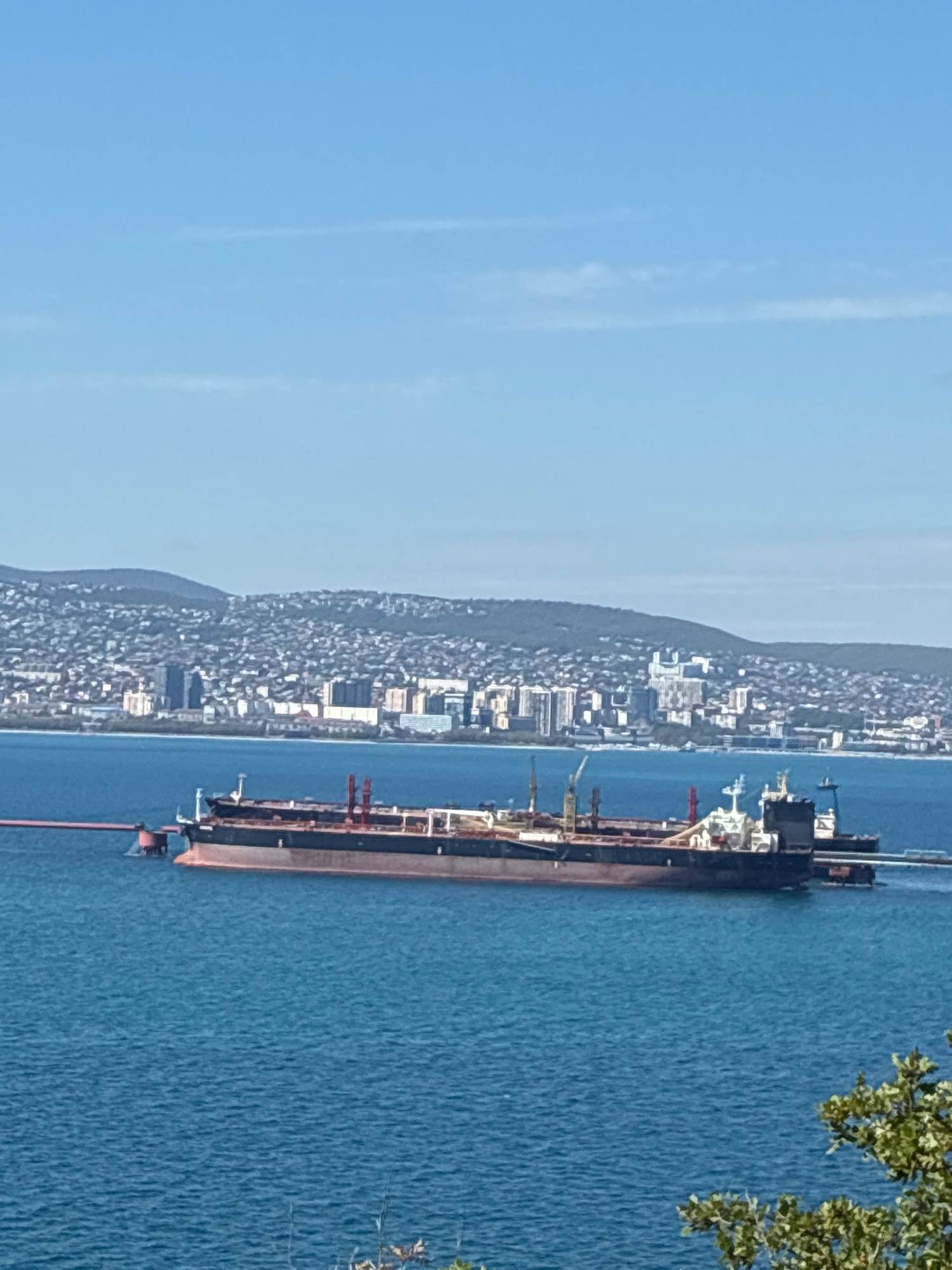 Two large cargo ships are anchored in the blue waters of the Black Sea, with the city of Novorossiysk, Russia, spread out on the hills in the background. The larger ship, a dark-colored tanker, is in the foreground, its bow facing left and extending towards a red loading arm. The tanker has a long deck with several red and white structures on it, likely cranes and loading equipment. A smaller ship is positioned behind the tanker to the right, also appearing to be a cargo vessel. The water is calm with small ripples, and the sky above is a clear, light blue with a faint wispy cloud. The city in the background features numerous buildings, ranging from low-rise structures to taller modern buildings, densely packed on the slopes of rolling green hills. The overall scene suggests a busy port environment.