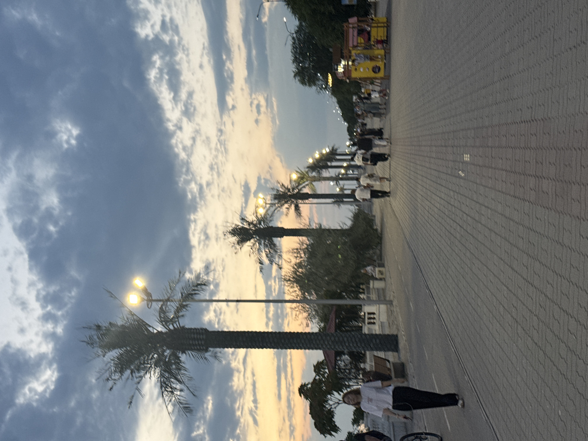 The image depicts a waterfront promenade in Aktau, Kazakhstan, during twilight. The sky is filled with dramatic clouds, transitioning from a warm yellow and orange near the horizon to a deep blue overhead, with streetlights illuminating the scene. Tall palm trees line the paved walkway, casting shadows and adding to the tropical atmosphere. Several people are visible, walking and gathered along the promenade. On the left side of the image, a woman in a white shirt and dark pants walks towards the foreground. Further down the promenade, a group of people stroll casually, some in groups, others individually. To the right, a vibrant yellow kiosk with colorful signs stands, suggesting a vendor or food stand. The ground is paved with rectangular bricks, creating a textured pathway. The overall scene suggests a leisurely evening atmosphere in a coastal city.