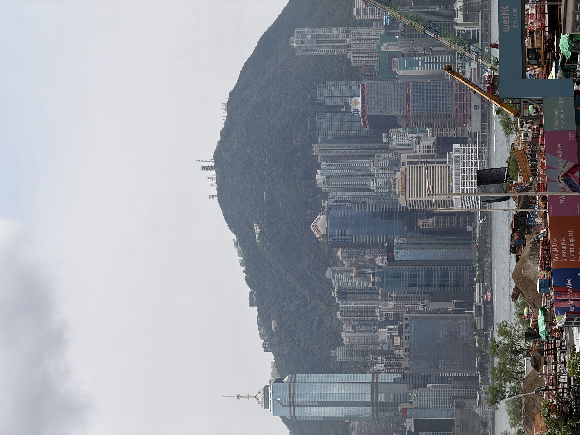 This is an image of the Hong Kong skyline, dominated by a large, verdant mountain in the background, known as Victoria Peak. The mountain is covered in lush green vegetation and dotted with residential buildings, and topped with numerous telecommunication towers. In the foreground, a dense collection of modern skyscrapers rises from the city. One particularly tall, slender glass skyscraper stands out on the left side of the image, distinguished by its distinctive spire. The city's buildings exhibit a variety of architectural styles and materials, with many featuring glass facades that reflect the overcast sky. In the lower right portion of the image, a construction site is visible, with scaffolding, cranes, piles of dirt, and heavy machinery. A large blue and teal structure with the word "westK" prominently displayed is part of the construction site signage. The body of water in front of the skyscrapers appears to be Victoria Harbour, with a few boats visible. The sky is mostly cloudy, contributing to a somewhat muted and diffused light.