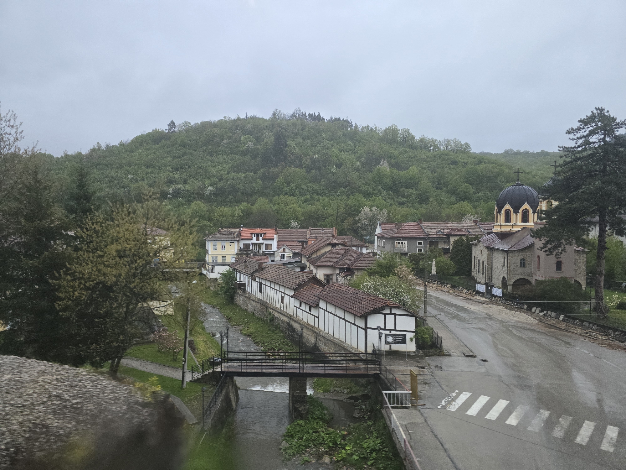 The image displays an elevated view of a quiet village or town situated in a valley, flanked by a lush, forested green hill under a uniformly overcast sky. The roads and surfaces appear wet, suggesting recent rainfall or light drizzle. No individuals are present in the scene, contributing to a tranquil and still atmosphere.
The settlement features various houses, many with characteristic red or brown tiled roofs. A notable row of white, half-timbered style buildings with dark roofs lines one bank of a winding river or stream that flows through the valley. A black metal bridge with railings spans the waterway, connecting the two sides.
Prominently featured on the right side of the image is an Orthodox church with a bright yellow facade, a dark brown roof, and a distinctive black dome crowned with a golden cross. A tall, dark green pine tree partially obscures the church on its right. A wet asphalt road, reflecting the overcast sky, runs past the church and parallel to the river. A white-striped pedestrian crossing is visible on the road.
On the road surface, near the pedestrian crossing, there is visible text with an arrow pointing right. This text reads "→ ГР. ТЕТЕВЕН", where "ГР." is an abbreviation for "град" (town/city) in Bulgarian. This indicates the location is the town of Teteven in Bulgaria. The time of day is daylight, but the heavy cloud cover results in soft, diffused lighting. The overall impression is one of peaceful serenity in a picturesque setting.