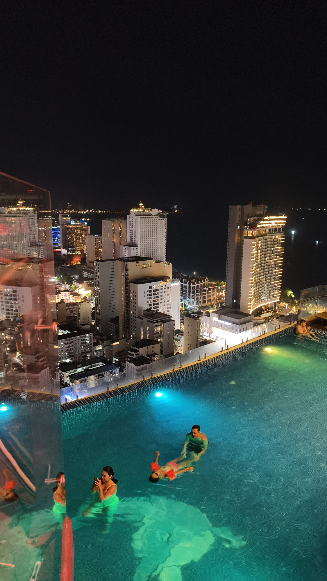 An aerial nighttime view captures a rooftop swimming pool overlooking a densely packed city skyline with numerous illuminated high-rise buildings. The pool, filled with turquoise water, is the foreground element, with several people interacting within it. In the lower left quadrant, two women are partially visible in the water, one holding a phone. To their right, a man is standing in the pool, looking towards a child who is floating on their back with arm floaties. In the middle ground, a man is seated in the pool on the right side, looking away from the camera. The city lights twinkle brightly, reflecting off the water's surface and the surrounding glass structures. The background shows a body of water, likely a bay or ocean, dotted with distant lights.