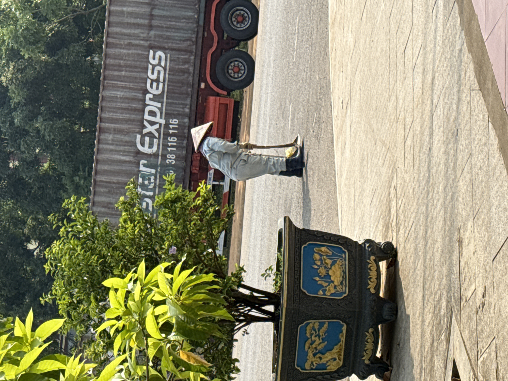 An outdoor scene in Bo Y, Vietnam shows a person wearing a conical Asian hat, light-colored work clothes, and black boots using a shovel to work on the side of a paved road. A large red and grey delivery truck with "Star Express" and the number 38 116 116 on its side is parked adjacent to the worker. A potted plant with bright green leaves and a dark-colored decorative pot with blue and gold designs is in the foreground. Behind the truck are trees. The scene is bathed in bright daylight.