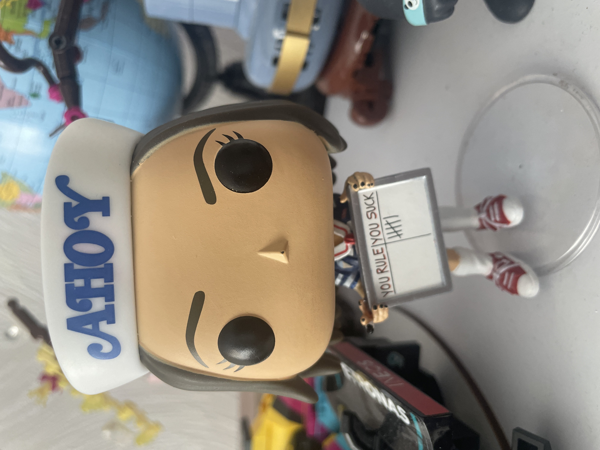 The image shows a close-up of a Funko Pop figurine of Robin Buckley from Stranger Things, wearing a white sailor hat with "AHOY" written in blue. She is holding a whiteboard with "YOU RULE" and "YOU SUCK" written on it, with tally marks under "YOU SUCK." The figurine is standing on a clear plastic base. In the background, out of focus, are various objects including a globe, what appears to be another toy, and part of a racing car model. The overall setting is likely an indoor display or shelf, given the arrangement of objects and the lighting. The image does not provide any information about the city of Brovary, Ukraine, or its specific location within it.