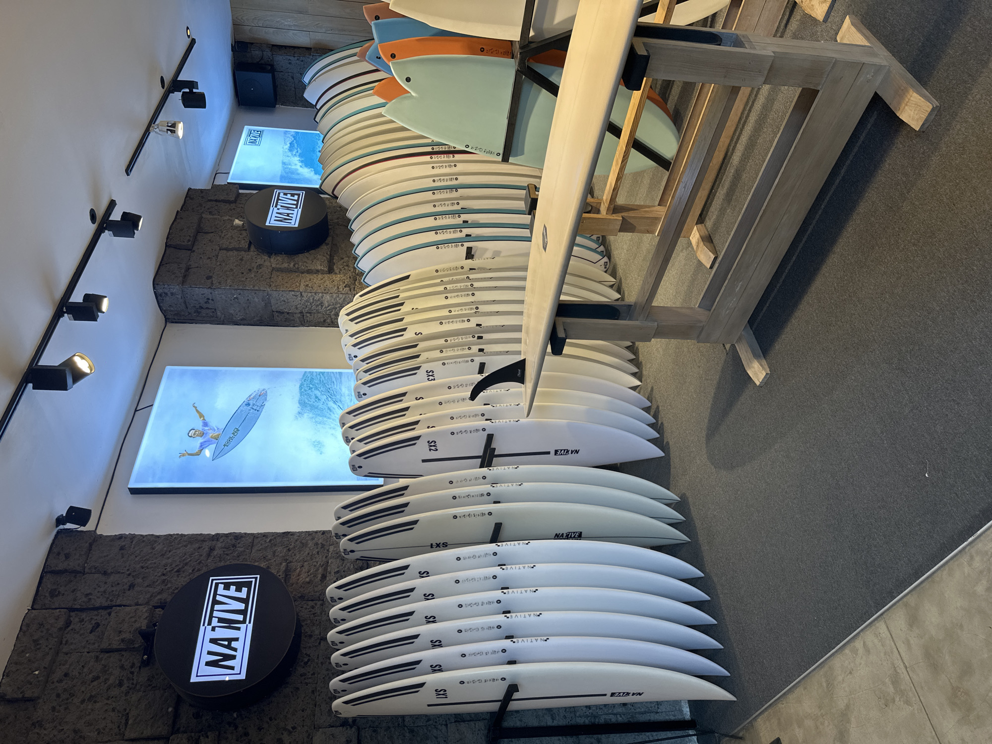 This is an interior shot of a retail store, likely a surf shop, given the number of surfboards on display. The boards are stacked vertically on a light-colored wooden rack, with several boards of varying sizes and designs. The surfboards closer to the bottom have white bodies with black accents, while the boards higher up have a variety of colors, including blue and orange.
The word "NATIVE" is prominently displayed on the boards, as well as on round black signs affixed to the stone wall. A sign also features a backlit image of a surfer in the water, emphasizing the theme. The environment includes a stone-textured wall, with track lighting on the ceiling. The floor is gray.
While the OCR suggests the location is North Kuta, Indonesia, this cannot be forensically confirmed by the image alone.