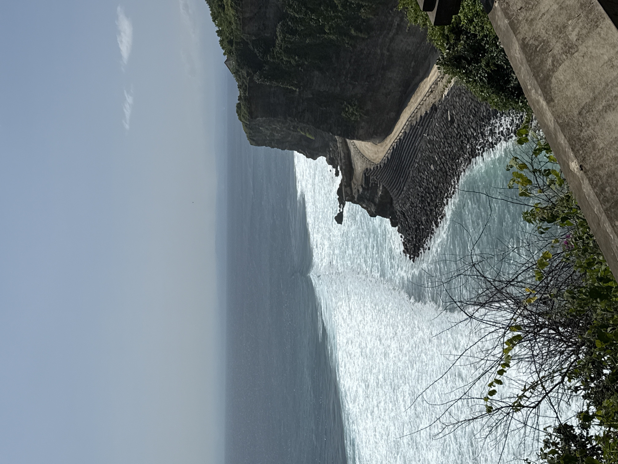 The image captures a coastal scene in Kuta Selatan, Indonesia. A large, rocky cliff face dominates the upper right, covered in dense vegetation. A pathway can be seen winding along the cliff's edge. Below, the ocean crashes against a dark, rocky shoreline, producing white foamy waves. The water extends into the distance, transitioning into a lighter, hazy blue. The sky above is a pale blue with a few wispy clouds. In the foreground, branches of trees and plants obscure part of the view, adding depth. A concrete structure is also visible in the foreground, partially obscuring the view.
