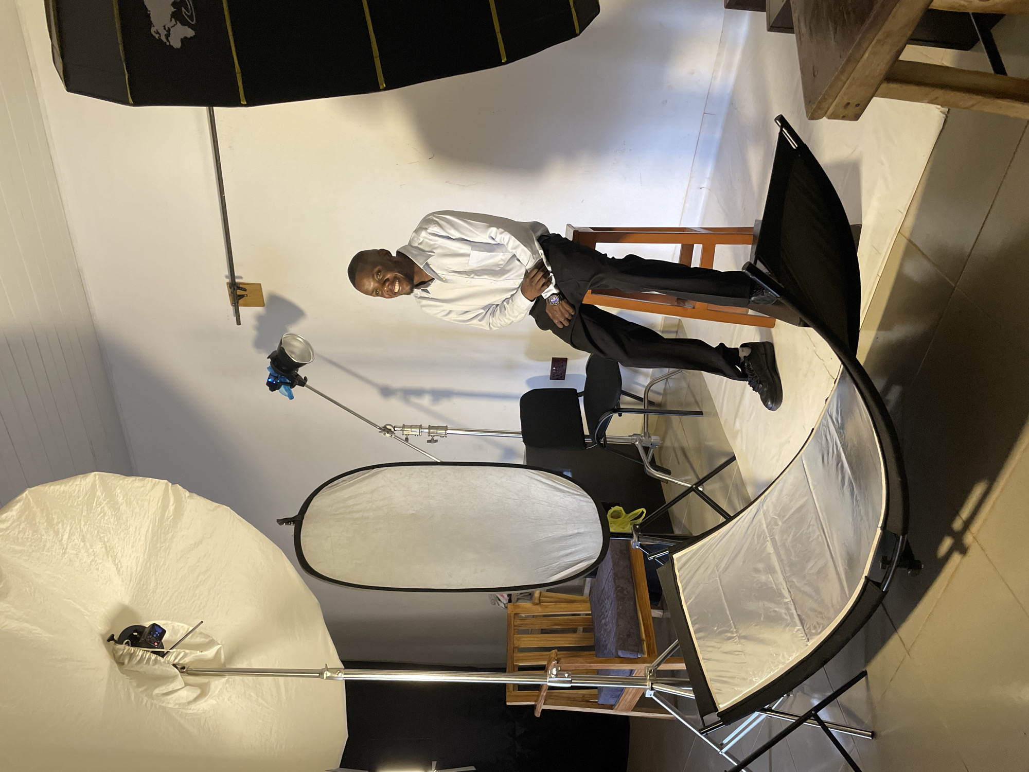 A Black man is seated on a wooden stool in what appears to be a photography studio. He is wearing a light blue button-down shirt and black pants. The man is smiling and looking to his left. The studio is equipped with various lighting equipment, including a large softbox on the left, a smaller softbox or modifier in the center, and another softbox on the right. There is also a reflector on a stand in the foreground. A wooden chair with a dark cushion is visible behind the man to his left, and a dark chair is to his right. The background is a white wall. The floor is tiled. The overall impression is a professional setup for portrait photography, possibly located in Durumi, Nigeria, given the context.