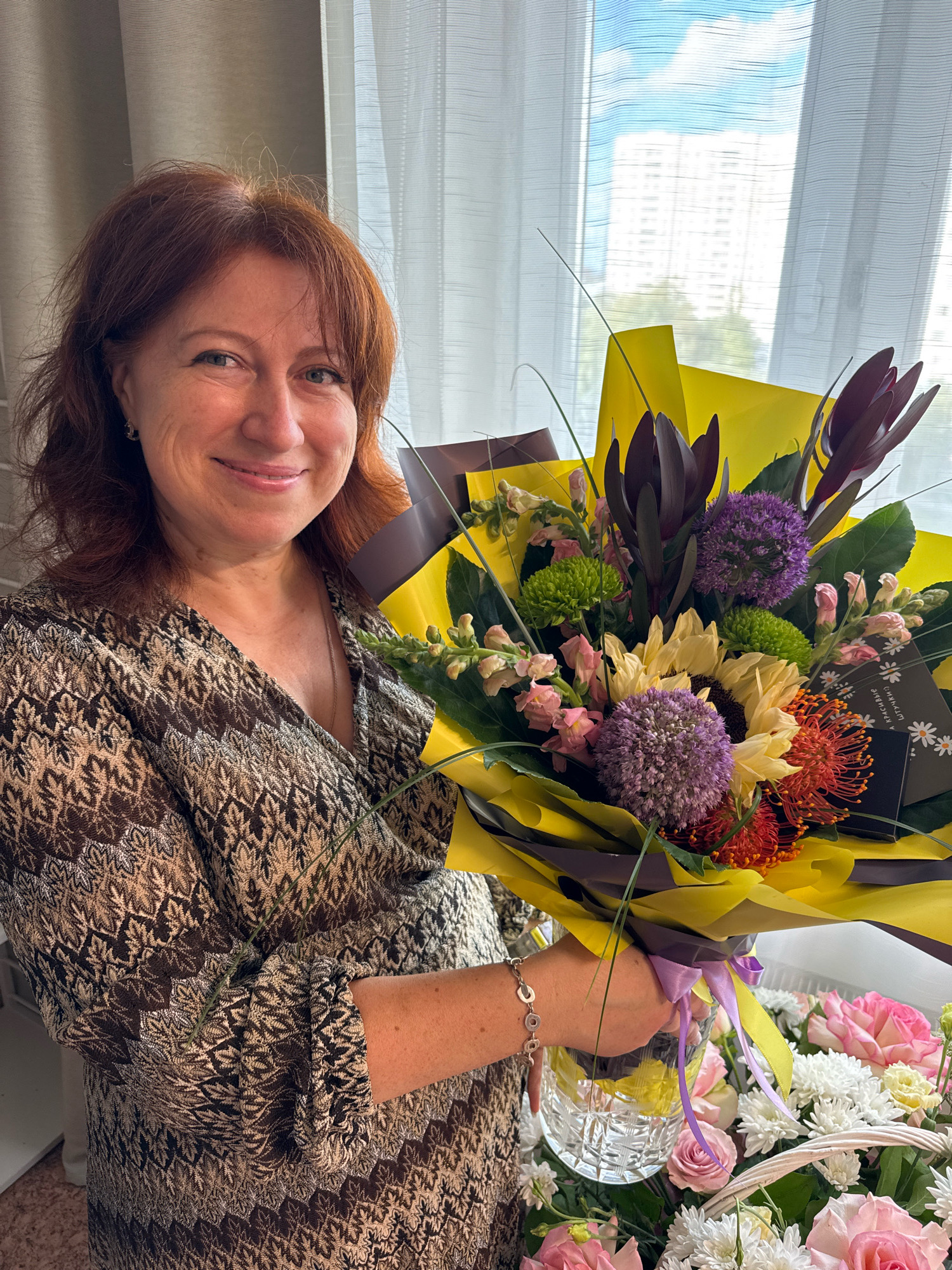 An exterior medium shot shows a fair-skinned Caucasian woman with auburn hair, smiling as she holds a large bouquet of flowers. She is wearing a brown and tan patterned blouse and a silver bracelet. The bouquet, which features sunflowers, purple alliums, green chrysanthemums, and red pincushion protea, is wrapped in yellow and brown paper. Behind her, sheer white curtains are drawn across a window that looks out onto a blurred city skyline and a bright blue sky. The woman is standing in what appears to be a residential building within the village of Zakharino, Russia. A basket of flowers is visible to her right.