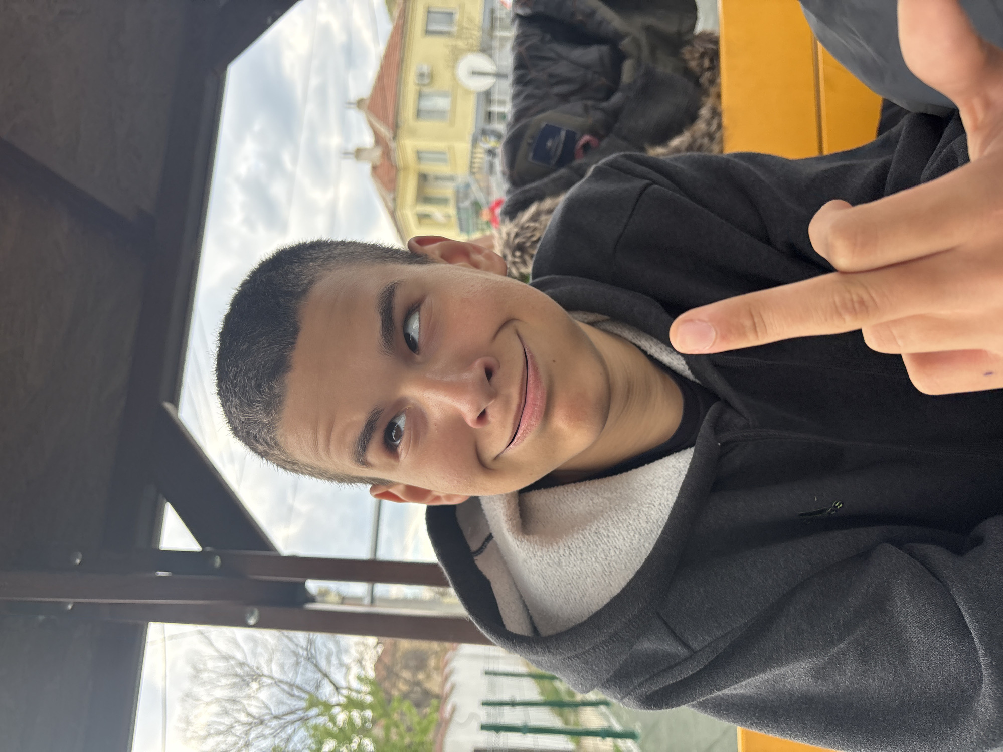 A young male with short, dark hair is indoors in a structure with wooden beams, possibly a gazebo or covered seating area, in an outdoor location of Unknown, Unknown. The subject is wearing a black hooded sweatshirt over a white undershirt. The person's index finger is extended straight out. In the background is a building, greenery, and the sky. The image appears to be taken from an oblique, upward angle.