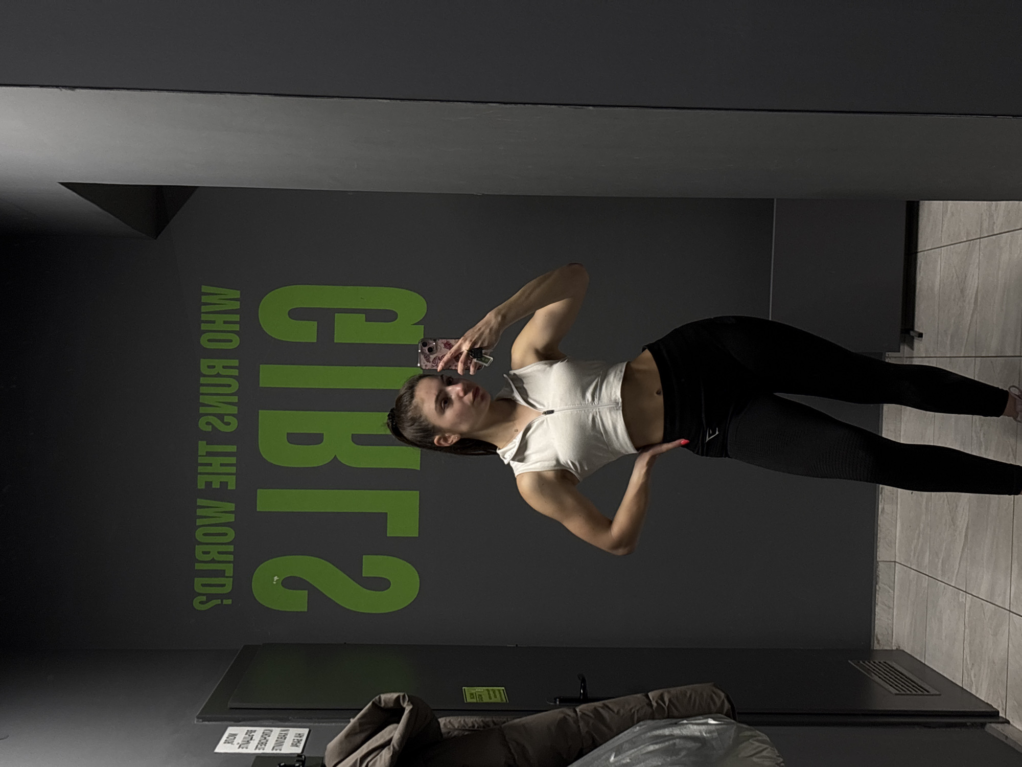 A young woman is captured in a full-body selfie, taken in what appears to be a gym or fitness facility. The image is presented rotated 90 degrees counter-clockwise. She stands in a pose, holding a smartphone with a patterned case in her left hand to take the photo, while her right hand rests on her hip. She has dark hair tied back in a ponytail and is wearing a white short-sleeved crop top with a zipper at the neckline, paired with black athletic leggings. Partially visible on her right foot are light-colored sneakers.
The background consists of dark grey walls. On the wall behind and to her left (when correctly oriented), large lime green text is visible, mirrored, which reads "WHO RUNS THE WORLD? GIRLS". The floor is covered with light-colored rectangular tiles. In the lower right corner of the image, there is a section of grey tiled wall and a dark vent on the floor. A dark object, possibly a bench or a neatly folded coat, is visible along the bottom edge of the frame. The lighting is indoor and artificial. The woman maintains a neutral expression as she poses.
