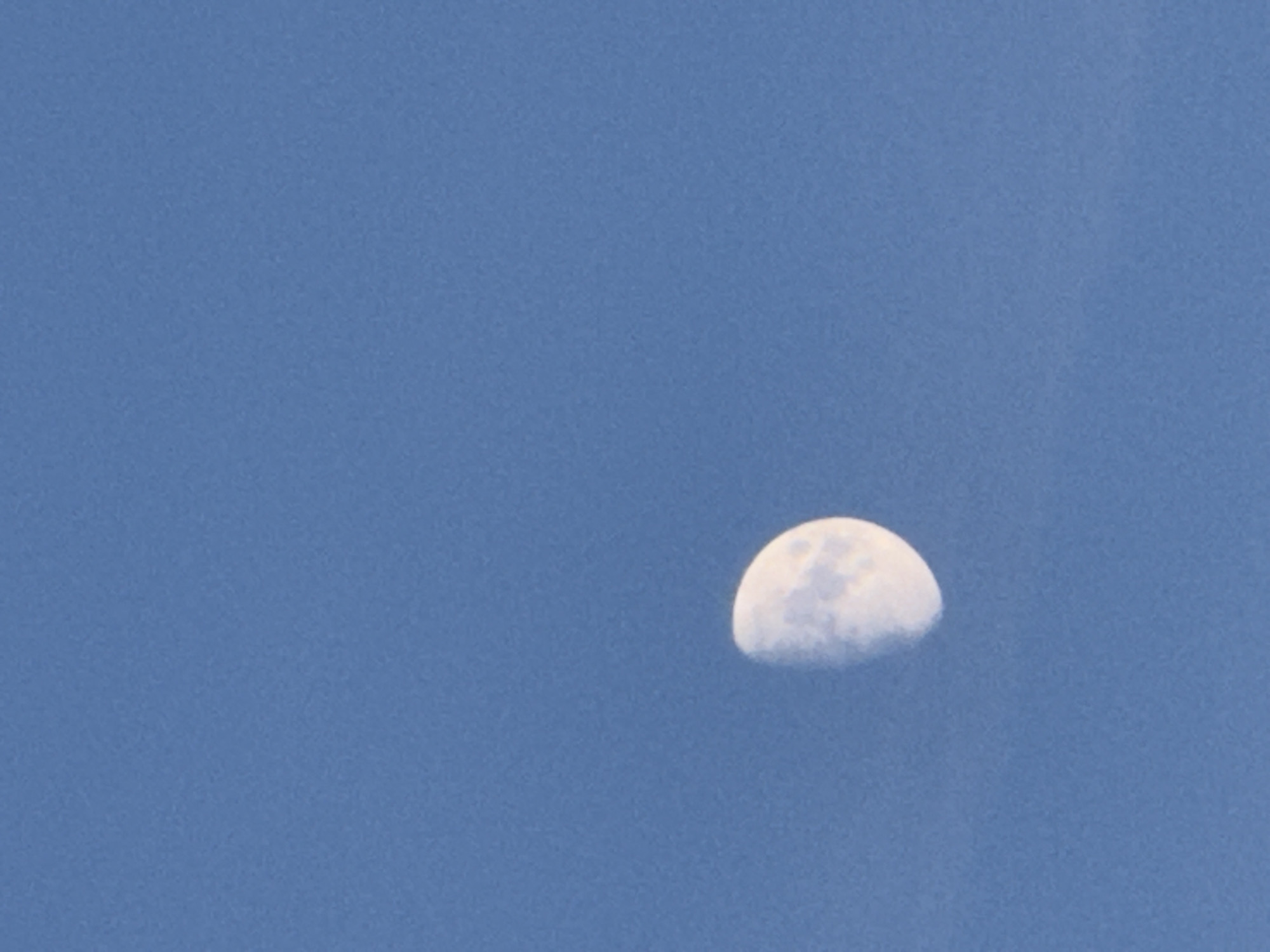 The image shows a partially illuminated moon against a clear blue sky. The moon is positioned in the lower left quadrant of the frame, showing its crescent phase. The sky is a uniform light blue, with no other celestial bodies or atmospheric phenomena visible. The photograph does not contain any elements that would suggest a specific location within the city of Ialoveni, Moldova.