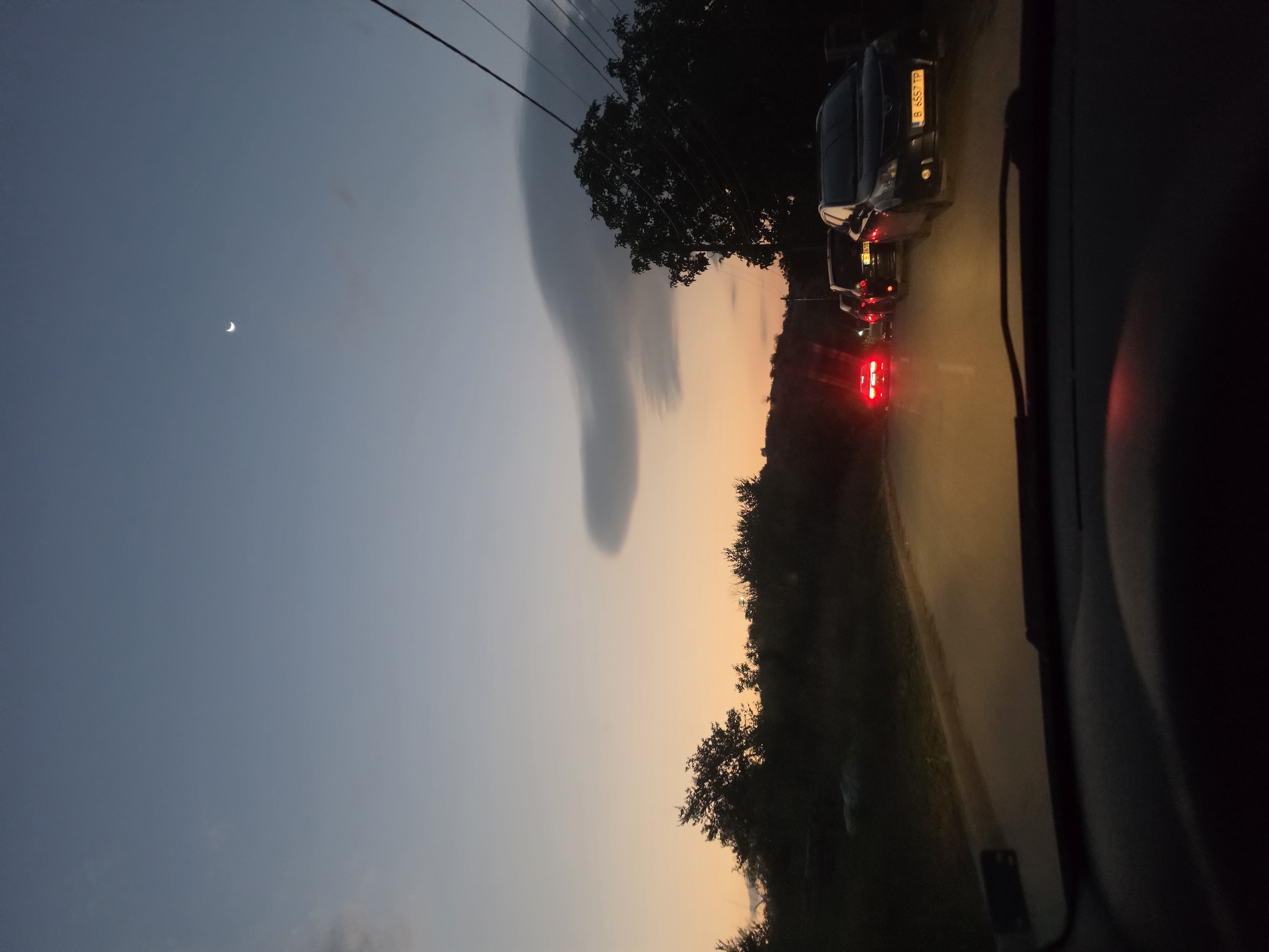 The image depicts a dusky scene viewed from inside a vehicle, presumably driving on a road near Strashimirovo, Bulgaria. The sky is a gradient of deep blue at the top, fading into a pale orange near the horizon, suggesting either sunrise or sunset. A crescent moon is visible in the upper part of the sky. A distinct, somewhat peculiar cloud formation is present in the mid-sky.
The road ahead is lined with trees and vegetation on either side, silhouetted against the dimming light. A line of cars is stopped or moving slowly on the asphalt road. The rear brake lights of the first car are illuminated red, and the brake lights of subsequent cars are also visible. The license plate of one of the cars reads "B 6557 TP". Overhead power lines are visible stretching across the frame, supported by poles that are mostly obscured by the trees. The foreground shows the interior of the vehicle from which the photograph is taken, with the dashboard and windshield wiper visible. The overall atmosphere is calm and quiet, typical of twilight driving.