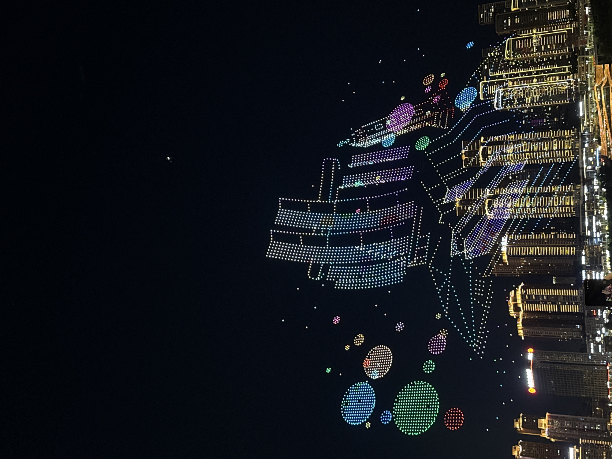 This is a nighttime cityscape view of Jiangbeicheng Subdistrict, China, dominated by a drone light show. Above a cluster of illuminated skyscrapers, thousands of drones form various shapes in the dark sky. Prominently displayed are circular formations of blue, green, and red lights, resembling bubbles or planets. To the right, a larger, more intricate formation of drones depicts what appears to be a ship or a stylized building, rendered in a spectrum of colors including white, blue, purple, and green. The buildings below are lit with numerous windows and vertical yellow lights outlining their structures. In the foreground, a dark expanse suggests a body of water or a lower landscape, with some blurred lights indicating further urban development. The overall scene captures a vibrant and technologically advanced urban night event.