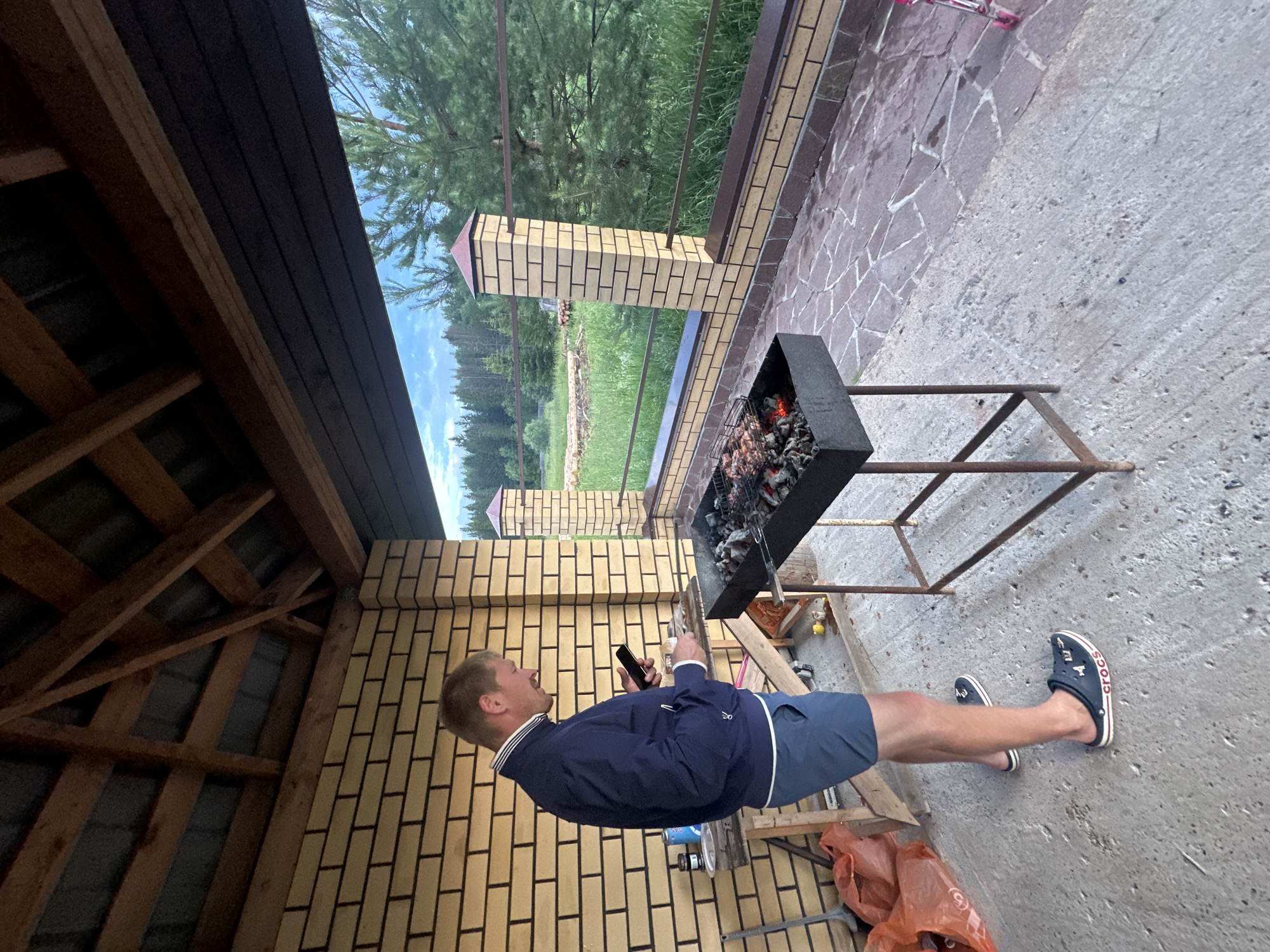 A man stands on a patio next to a barbecue grill, holding a mobile phone and a small white object. He is wearing a dark blue jacket, blue shorts, and dark blue Crocs. The barbecue grill contains burning coals and meat on a rack. The patio is paved with stone and concrete. Behind the patio is a brick wall and a wooden roof structure. In the background, there is a lawn, trees, and a forest. The scene appears to be outdoors, possibly in a backyard or a rural area. The sky is cloudy.