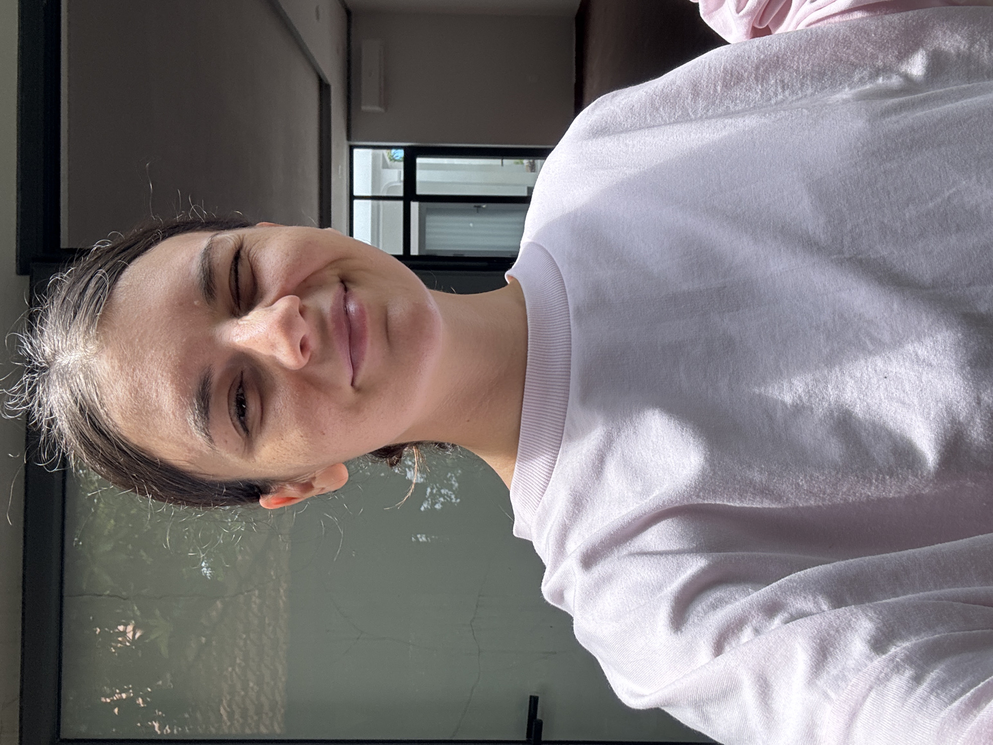 In an outdoor setting in Mũi Né, Vietnam, a young woman with dark hair is taking a selfie. She is looking at the camera and winking, with a slight smile on her face. She is wearing a light pink long-sleeved sweatshirt. The background shows a glass door and a building interior with dark flooring and a white ceiling. Sunlight is casting shadows on her face and sweatshirt.