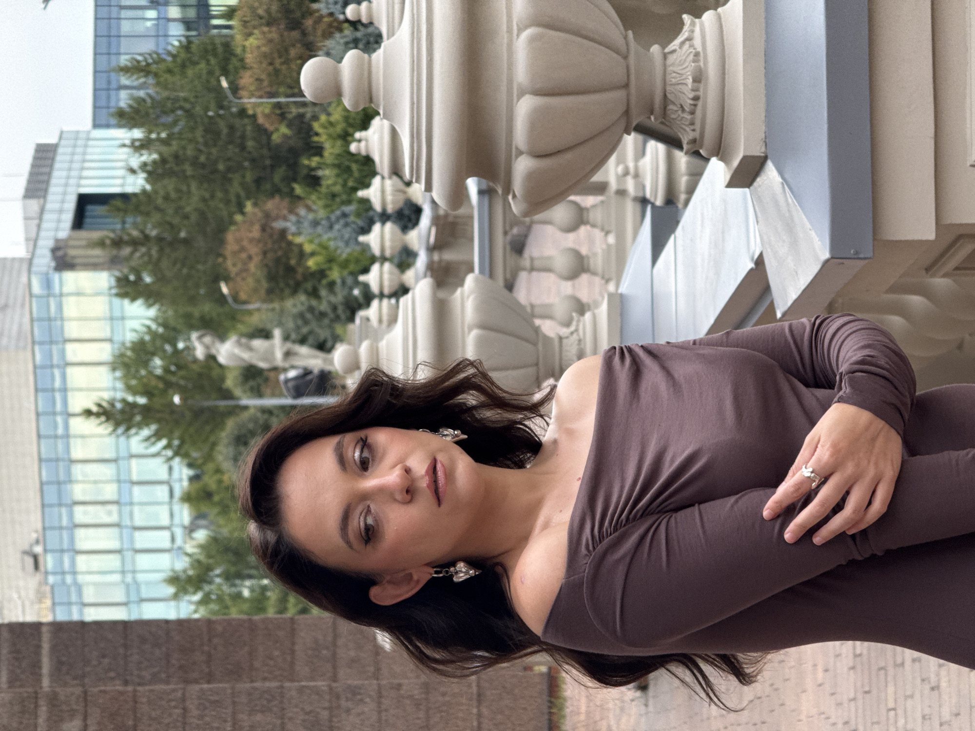 A medium shot captures a woman with long, dark, wavy hair, wearing an off-the-shoulder, long-sleeved brown top. She has heart-shaped earrings and a ring on her left hand. Her gaze is directed to her left, with her left arm bent, resting across her torso. She stands on a paved area, with ornate white balustrades and urns in the foreground. Behind her, a modern building with glass windows is visible, surrounded by trees. The scene is outdoors, suggesting a public space in the city of Kazan, Russia.