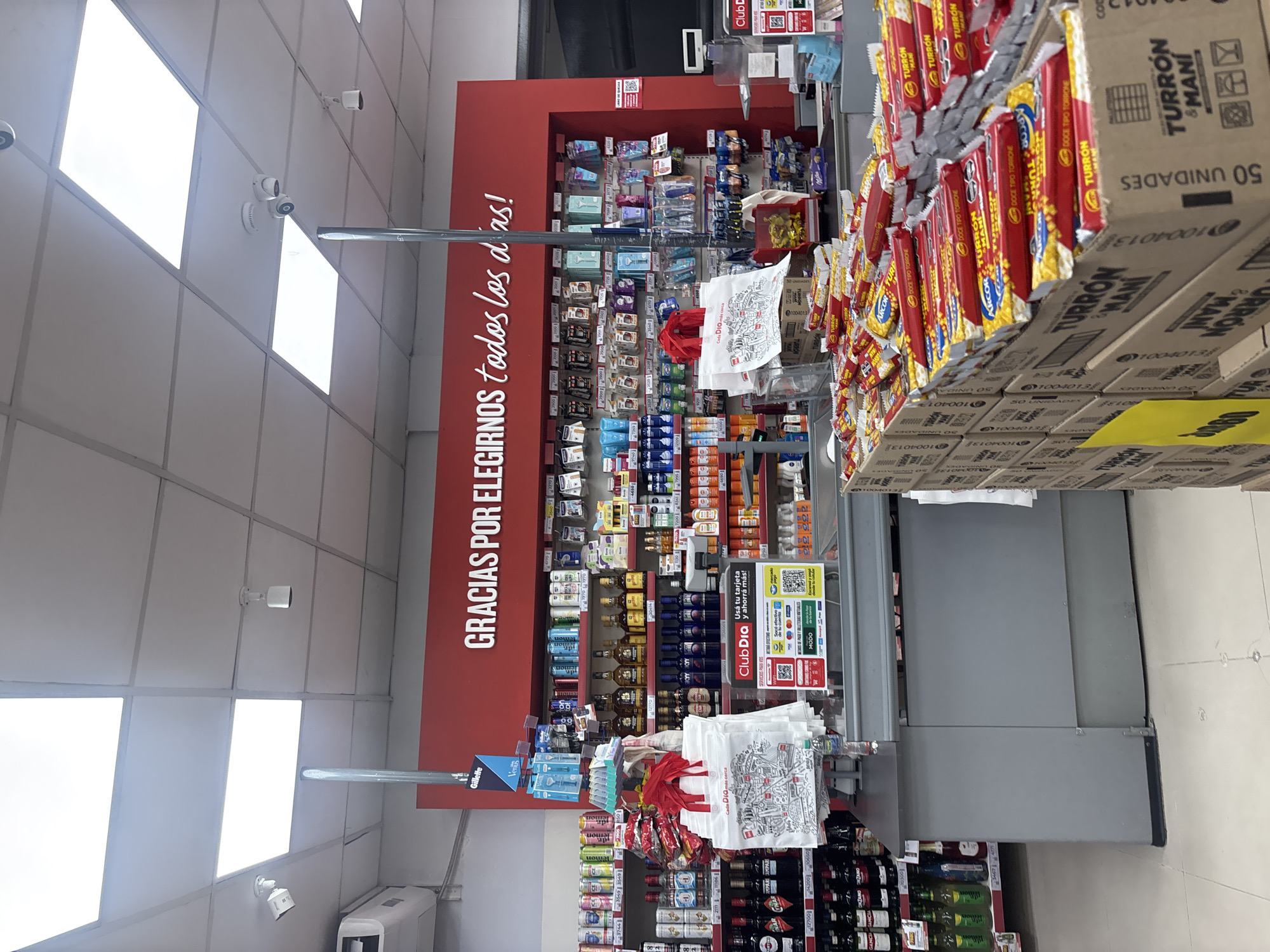 This is an interior shot of a retail store, likely a supermarket or convenience store, in Buenos Aires, Argentina. The store features aisles of products, with a checkout lane visible. Shelves are stocked with various items, including packaged candies, beverages, and general merchandise. The shelves are well-organized. The red wall displays the phrase "GRACIAS POR ELEGIRNOS todos los dias!", which translates to "THANK YOU FOR CHOOSING US every day!" indicating a customer-centric message from the store. The cashier area is visible, with boxes of "Turron" stacked high. Several surveillance cameras are visible in the ceiling. The products are standard consumer goods.