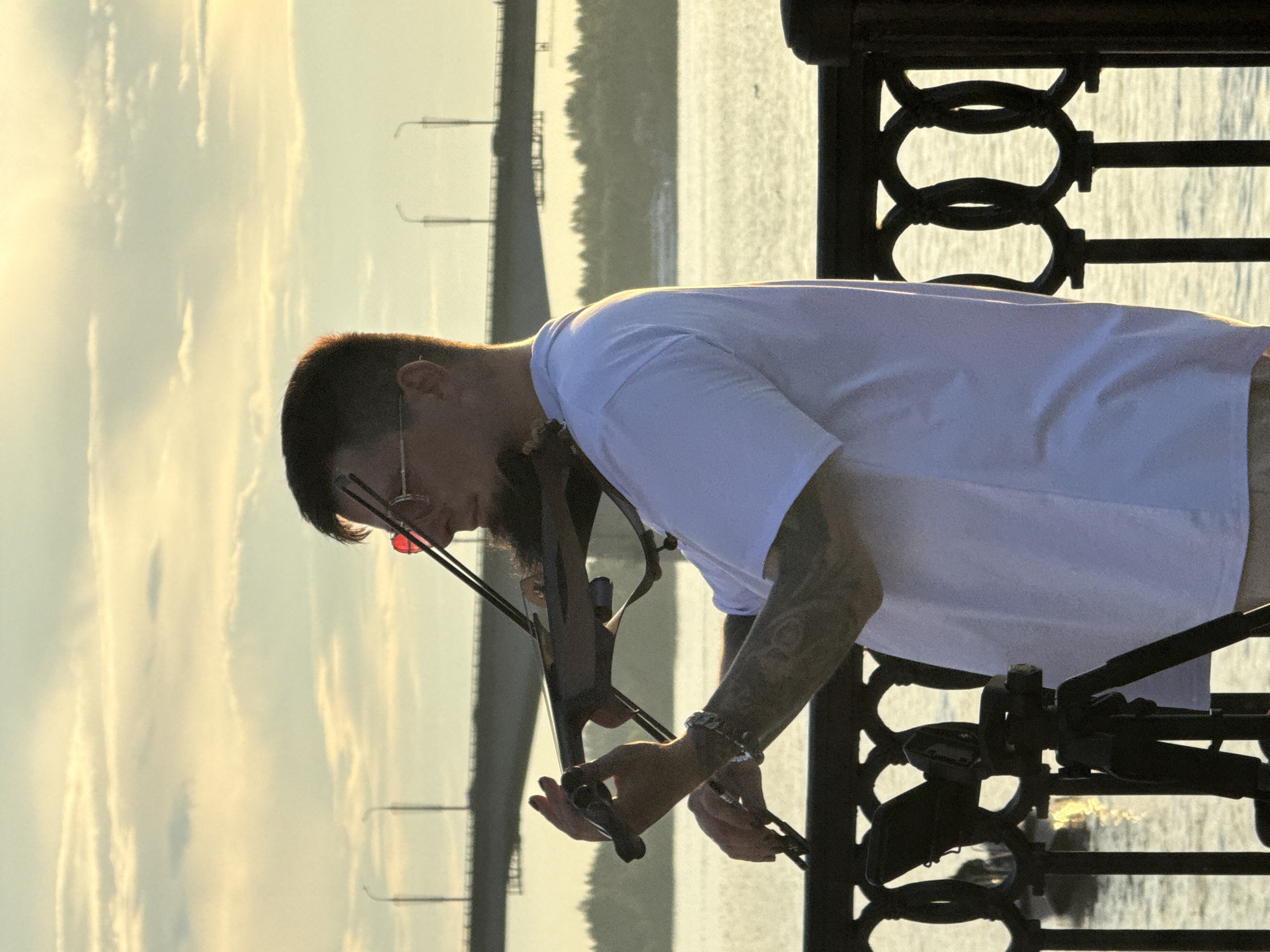 A man with short dark hair, a beard, and sunglasses with red lenses plays a violin outdoors at sunset. He is wearing a white t-shirt and beige shorts, and a silver bracelet on his left wrist. His forearms are covered in tattoos. He is standing on a walkway with a black metal railing, in front of a body of water. In the background, a large bridge spans the water, with trees and buildings visible on the far bank. The sky is filled with soft, orange and yellow clouds. A tripod with a camera is visible in the foreground to the left of the man. This scene appears to be located on the embankment of the Kama River in the city of Perm, Russia, likely near the Kamges hydroelectric station bridge.