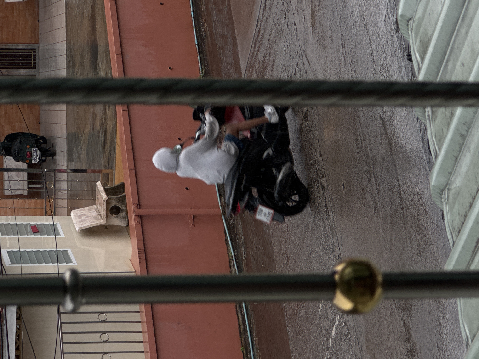 A person wearing a light-colored hooded jacket and dark shorts rides a dark-colored scooter on a wet, asphalt road. The road surface shows ripples and puddles from rainfall. The scooter has a license plate that is partially visible. In the background, a red wall runs along the side of a building. Further back, there are barred windows and a section of brick wall. A dark scooter is parked in front of a building with a license plate that reads "H 2655". A stone bench is visible next to a wall with louvred windows. The scene appears to be outdoors, with visible signs of rain.