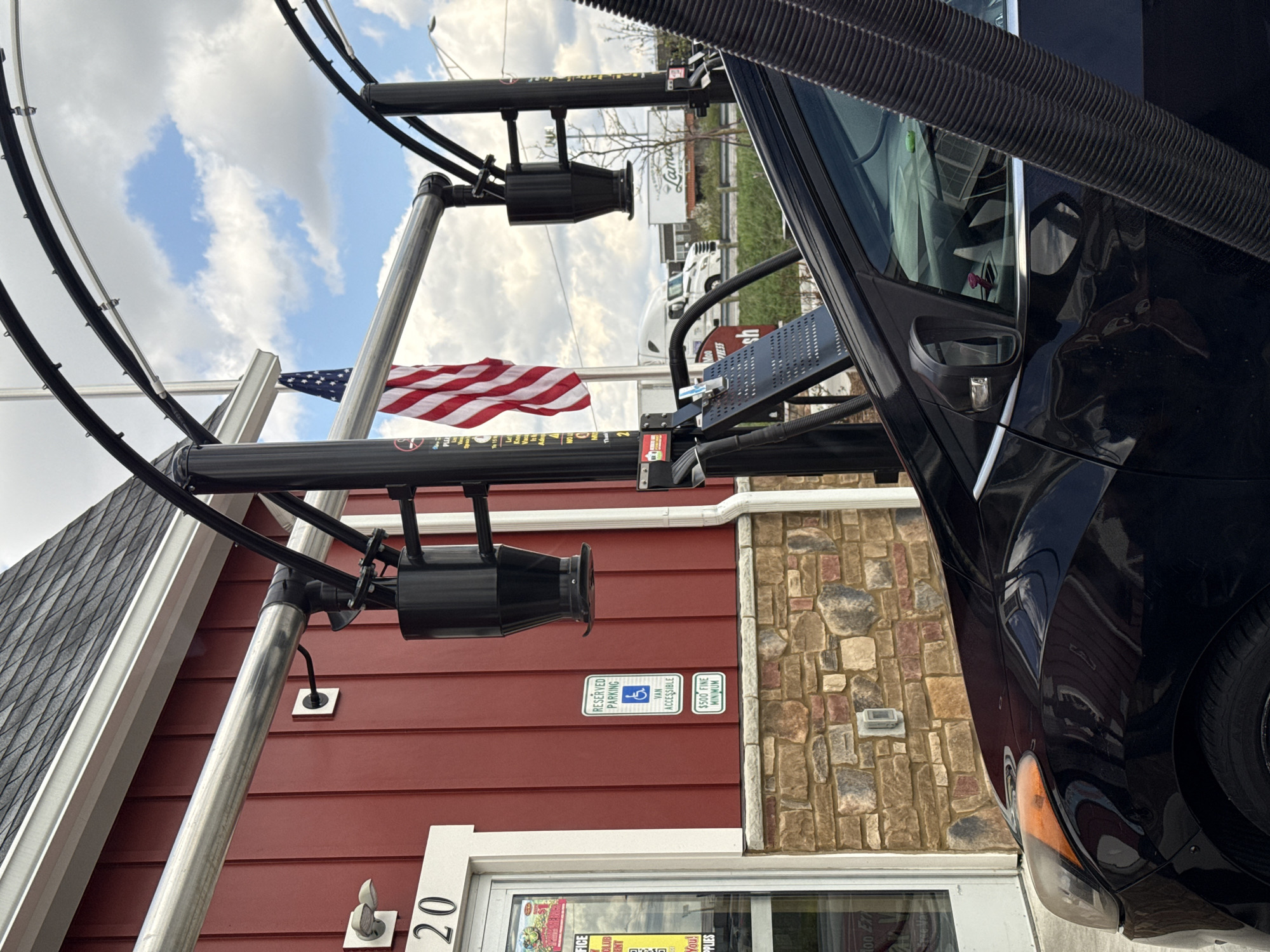 The image depicts a car wash entrance at 20 Westmoor, Columbus, OH 43204, as determined by the signage in the window and the street address "20" above the door. A black car is partially visible on the right side of the frame, suggesting it's about to enter the car wash. The washing apparatus, with cylindrical sprayers, is mounted on a silver and black metal structure. The building has a red exterior with white trim and a section of stonework. A handicapped parking sign is visible on the wall. An American flag waves in the background.