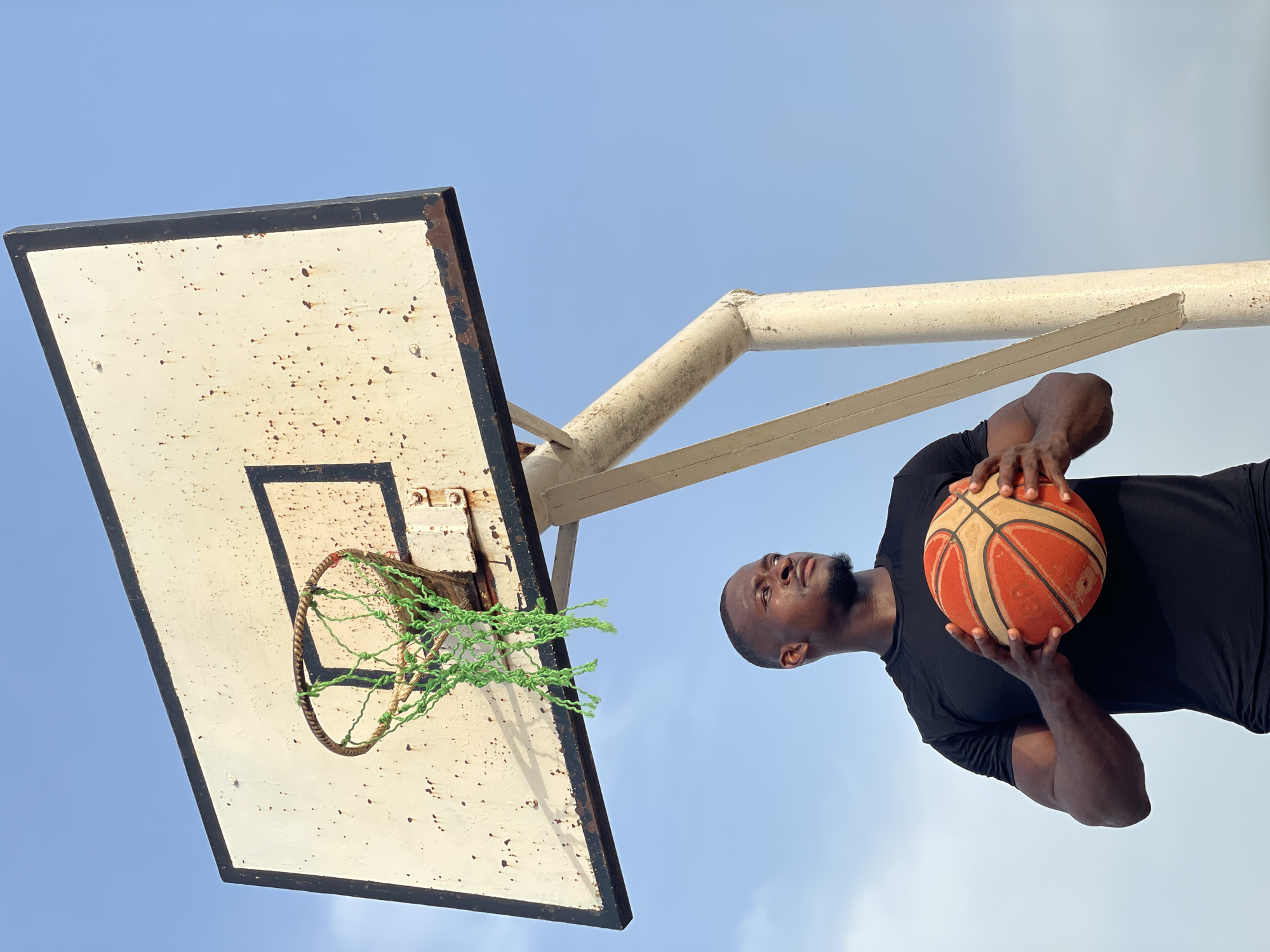 The image depicts a man holding a basketball, standing in front of a weathered basketball hoop in Akwanga, Nigeria. The hoop has a white backboard with a black border and a rusty metal ring with a green rope net. The man is dark-skinned, wearing a black shirt, and looking upwards. The background is a clear, light blue sky. The basketball is orange with black lines.