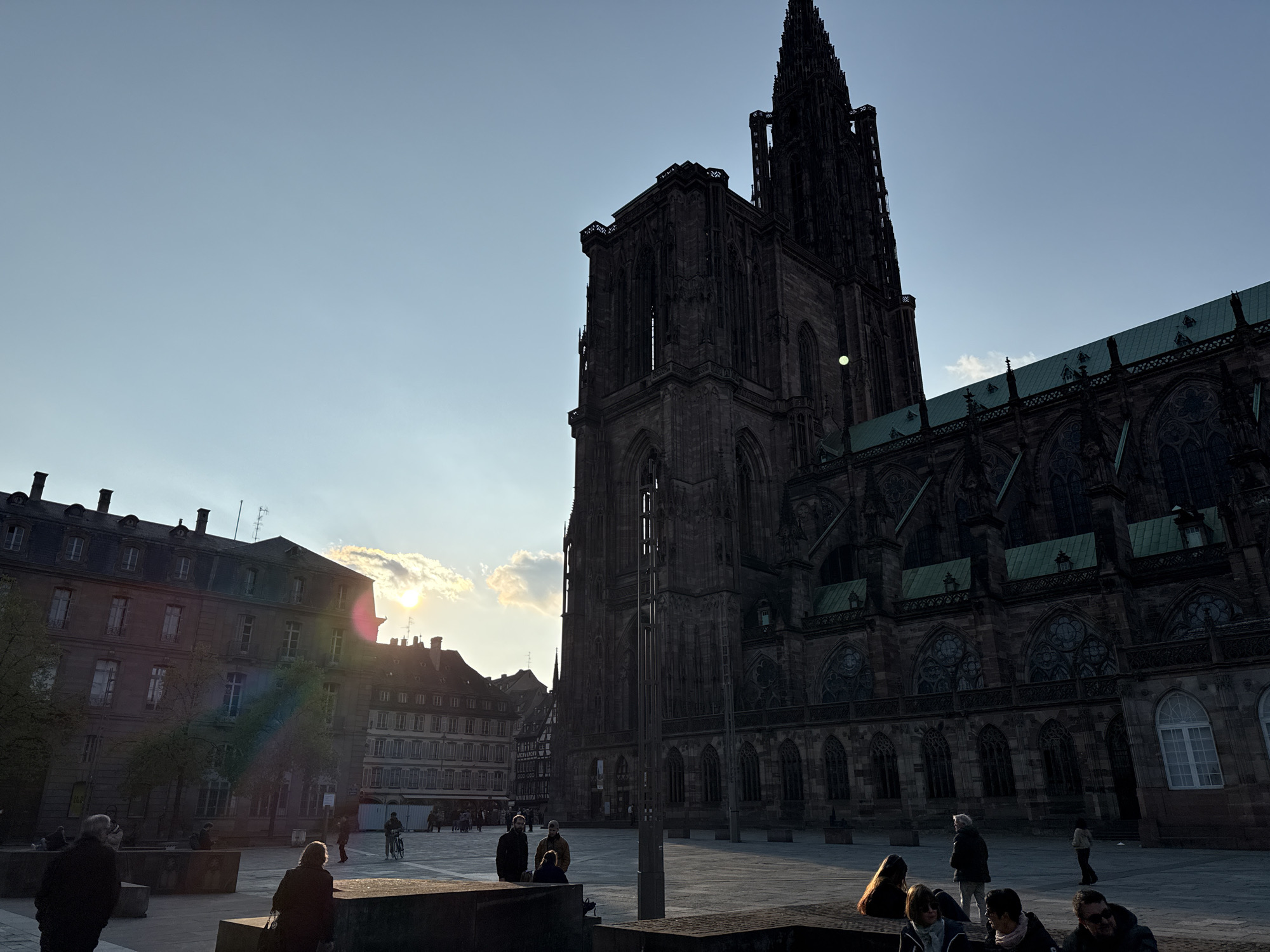 The image shows a cityscape with a large Gothic cathedral dominating the scene. This is the Strasbourg Cathedral, located in Strasbourg, France. The cathedral is in the center-right of the image, silhouetted against the sky. To the left of the cathedral, several buildings can be seen, with the sun positioned between them, creating a bright lens flare. In the foreground, a public square is visible, with several people sitting and standing around. Some people appear to be interacting in small groups. The sky is clear and blue, with some clouds near the horizon.