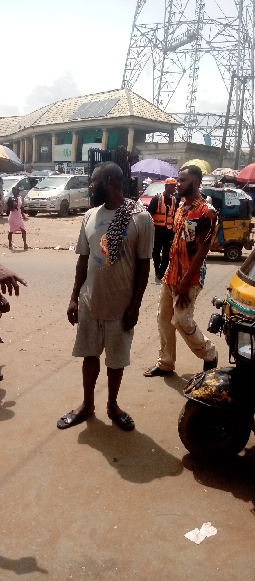 On a sunlit street in Onitsha, Nigeria, two men are the focal point. One man, wearing a grey t-shirt, grey shorts, and black sandals, stands with a patterned cloth draped over his shoulder. The second man, walking past him, is clad in a vibrant, patterned short-sleeved shirt and tan pants, also wearing sandals. Behind them, a man in an orange vest and cap is visible. A yellow auto rickshaw ("keke") is partially in the frame, close to the second man. Other vehicles and pedestrians are visible in the background, including a woman in a pink dress. Commercial buildings line the street, some with solar panels on the roof. A tall telecommunications tower stands in the background against a bright sky. The street is unpaved and dusty.