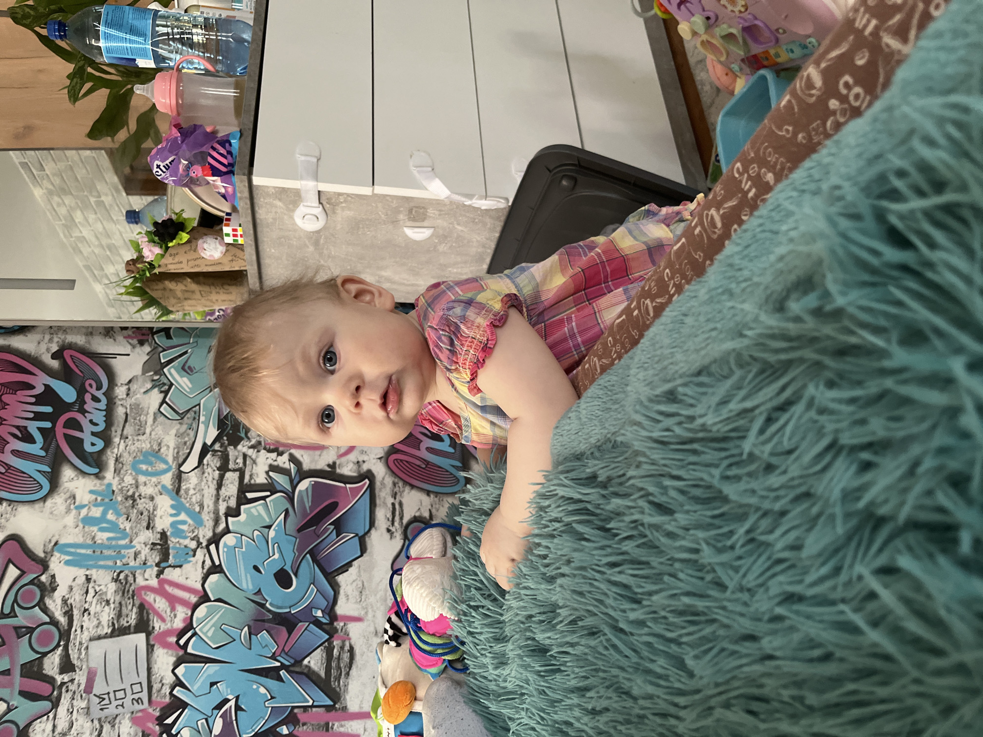A close-up shot displays a young child with fair skin and light hair, lying on a fluffy, light-blue material with a graffiti-style design wall in the background. The child is partially dressed in a plaid shirt with red, yellow, and purple colors, holding onto the fluffy material. The surrounding environment features a collection of objects, including bottles, toys, and storage units, suggesting an indoor setting. There is no mention of Vesyoloye, Russia.