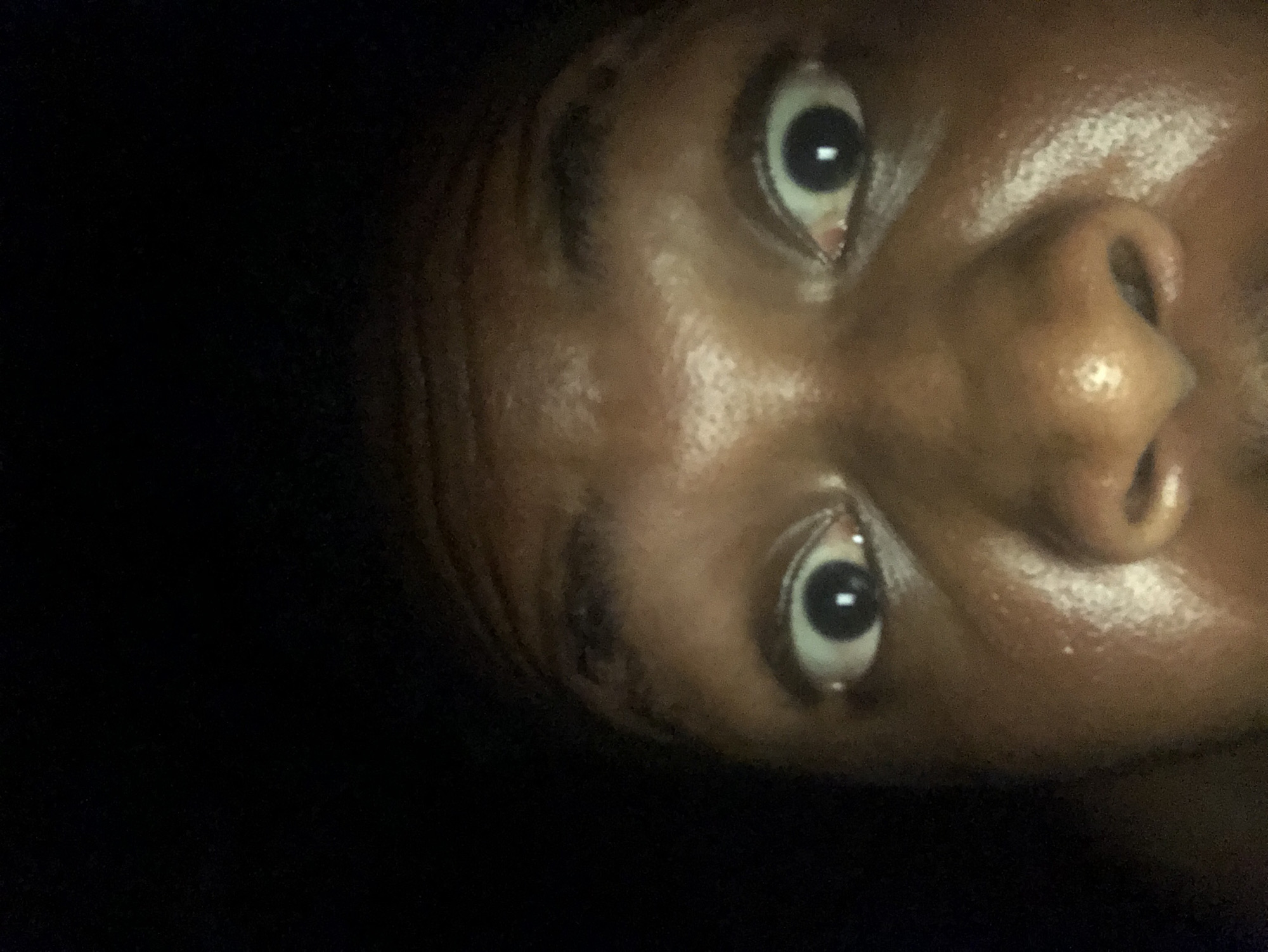 A close-up, angled shot of a person's face. The person has dark skin, dark eyes, and a visible nose. The background is dark, suggesting low lighting conditions. The image appears to be taken at an upward angle.
