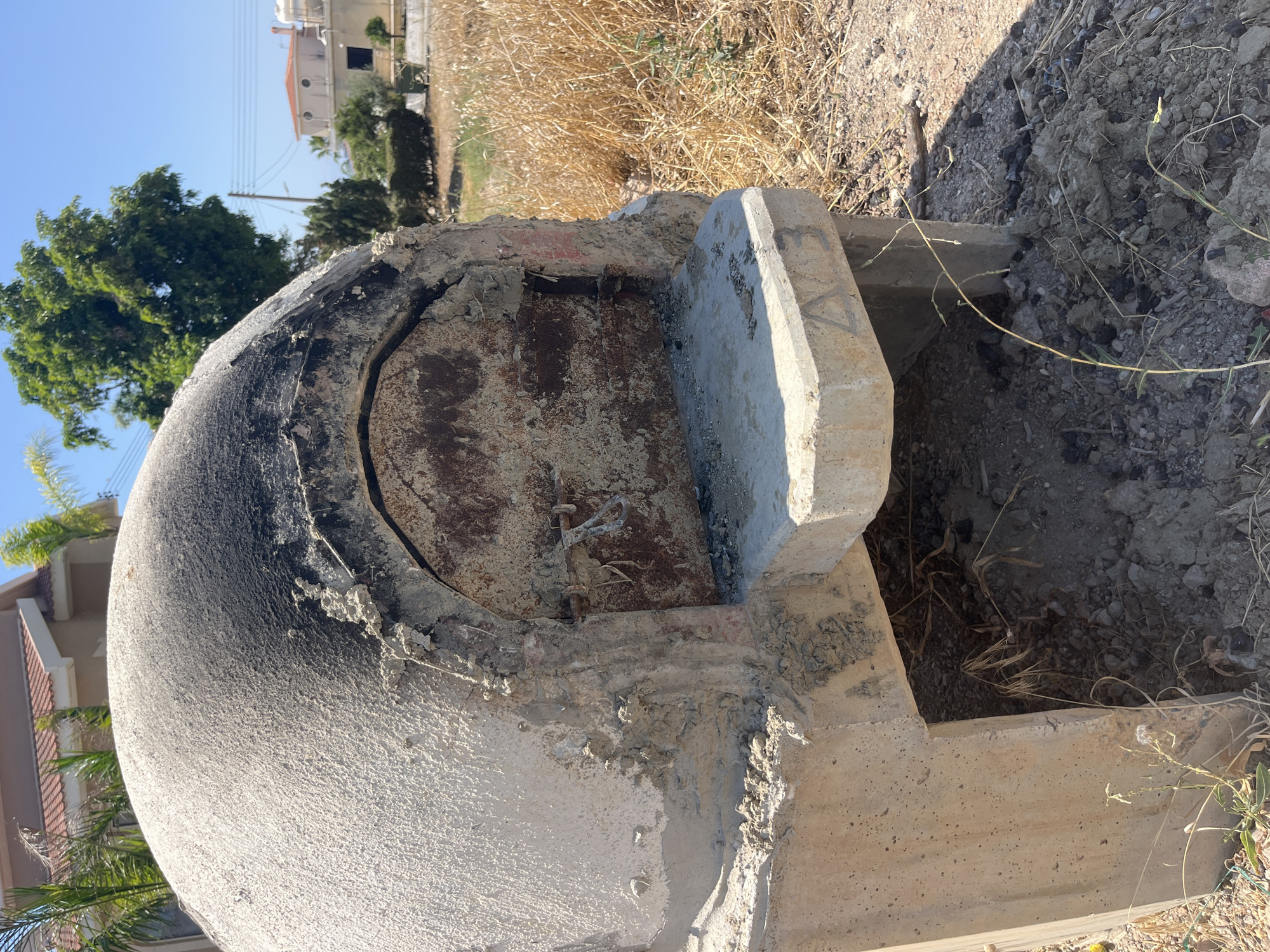 A rustic, dome-shaped oven made of concrete and plaster sits in an outdoor environment. The oven is supported by a concrete base with an opening underneath. The dome has a weathered appearance, with areas of soot near the top and around the opening. A metal door, rusted and grimy, covers the oven's mouth. The ground around the oven is covered in dry grass, dirt, and small stones. In the background, there are buildings with terracotta roofs and lush green trees under a clear blue sky. The location is identified as Moni Community, Cyprus, suggesting a possibly rural or semi-rural setting with residential structures nearby.