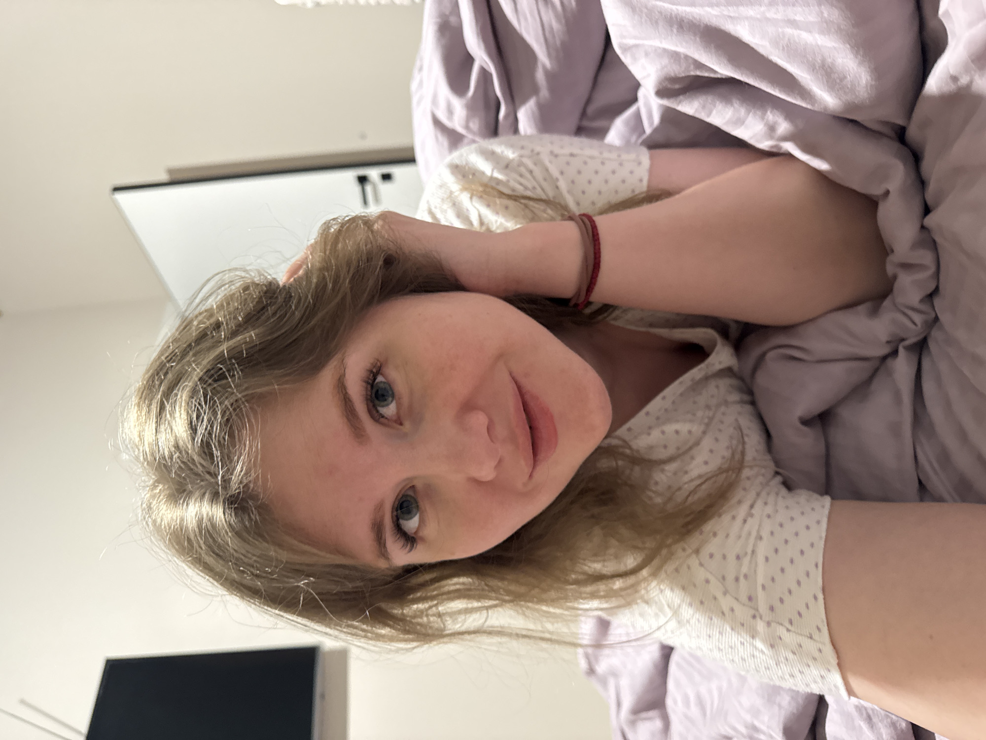 The image is a close-up portrait of a young woman lying in bed, looking directly at the camera with a gentle, relaxed expression and a subtle smile. She has long, light brown or blonde hair that frames her face and distinct blue eyes. Her complexion is fair, with a scattering of visible freckles. She is dressed in a light-colored top, likely pajamas, featuring a faint polka-dot pattern. On her right wrist, she wears two thin bracelets, one red and one dark-colored.
She is partially covered by a soft, light lavender-colored blanket or duvet, which is casually draped around her. Her left arm is bent, with her hand resting near her head and lightly touching her hair. The setting appears to be an indoor space, most likely a bedroom, with a softly blurred background. Elements visible in the background include a plain white wall and a light-colored surface, possibly part of a headboard or furniture, in the upper area of the frame. A dark, rectangular object, possibly a screen or monitor, is visible in the lower left.
The lighting is soft and even, creating a calm and intimate atmosphere, consistent with a bedroom setting during daytime or early morning/evening. There is no visible text in the image. The location context provided (Gornyi, Russia) is not visually discernible from the image itself.