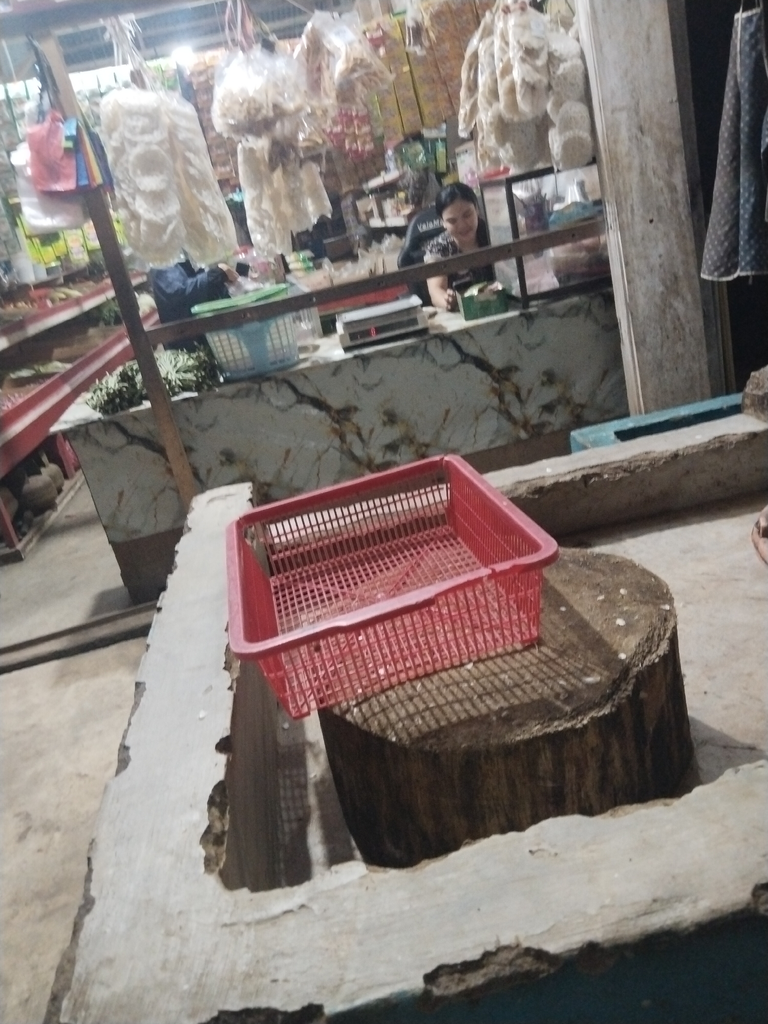 The image depicts an indoor market stall in Suka Ramai, Indonesia. A woman, likely a vendor, is visible behind a counter, interacting with a customer or arranging items. The stall is stocked with various goods, including what appear to be dried snacks or food items packaged in plastic, hanging from the ceiling. In the foreground, a bright red plastic basket sits on a wooden chopping block, casting a grid-like shadow. The environment suggests a bustling local marketplace with a mix of goods and everyday activity.