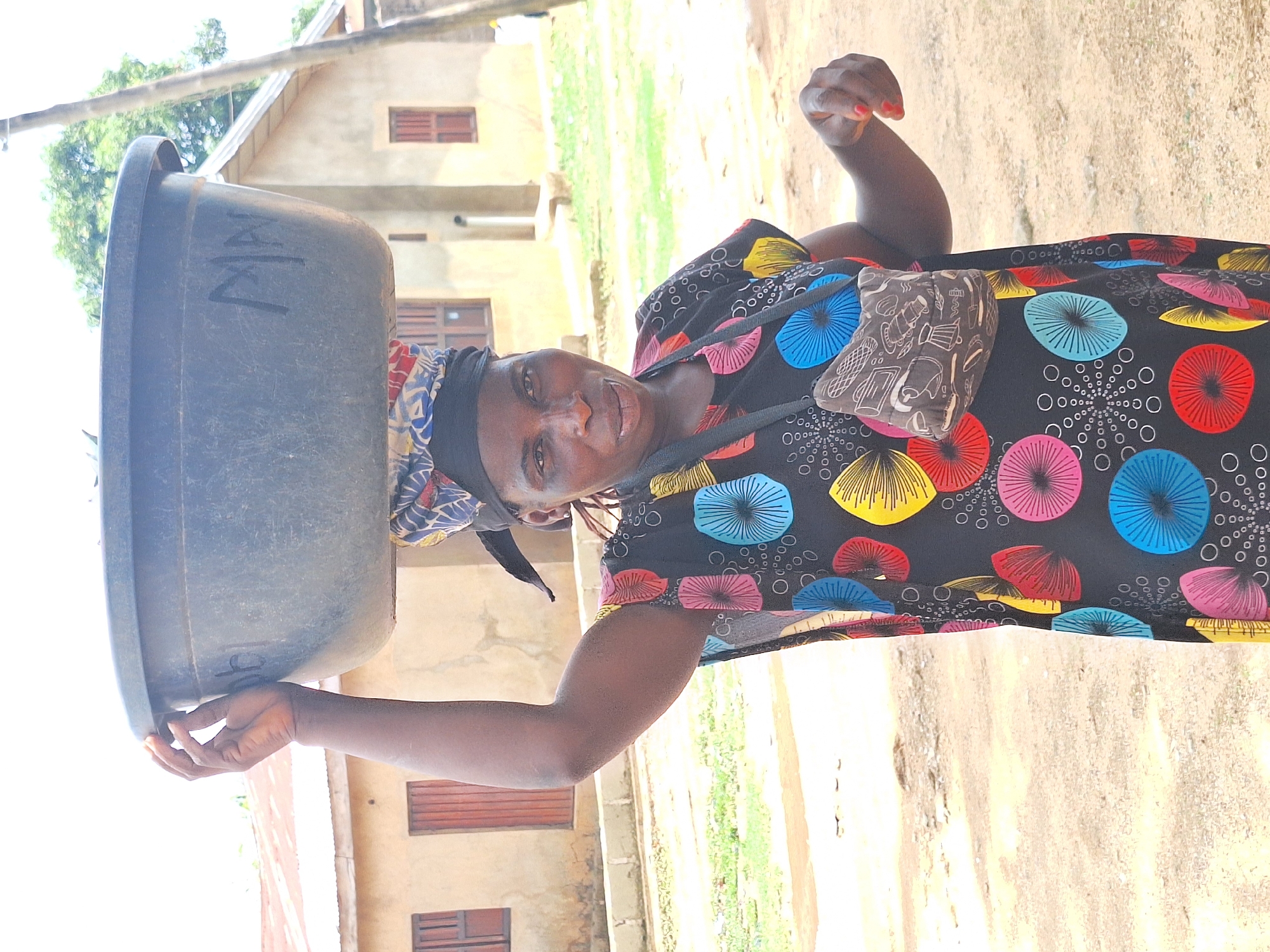 The image shows a woman standing in Sabon Tasha, Nigeria, with a large, dark gray tub balanced on her head. The tub has the letters "MZ" written on its side in white. The woman is wearing a colorful patterned dress with a black background and designs featuring circles and radiating lines in blue, yellow, pink, and red. She has a small bag slung across her body. A building with brown shutters is visible in the background, and the ground appears to be a dirt or gravel path. The lighting suggests it is daytime.