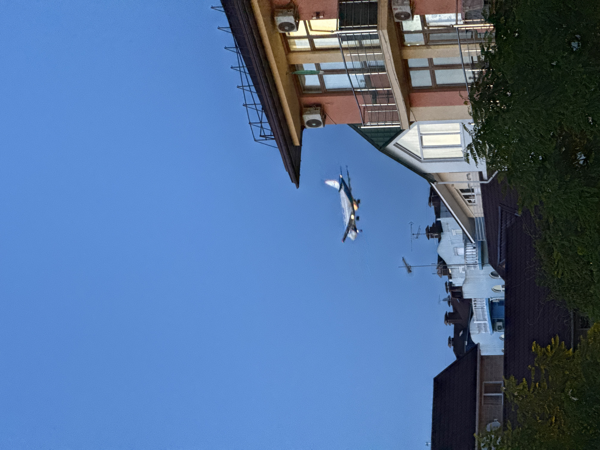 The image, rotated ninety degrees clockwise, depicts an urban scene under a clear, deep blue sky, likely at dusk or early evening. The prominent subject in the sky is an elongated, metallic-looking aircraft, possibly a jet or drone, with discernible wings and a tail section, appearing to be in flight. Below it, two smaller, very faint, white elongated objects are also visible in the sky.
On the left side of the image, a multi-story building with a light-colored facade (possibly peach or light orange) and a dark overhang is visible. This building features multiple rectangular windows, some reflecting light, and at least two external air conditioning units. A metal framework or scaffolding is attached to its upper side.
To the right, another building, lighter in color (white or light blue), is partially visible, showing several windows, balconies, and possibly antennas on its roofline. A darker building or section of a building is further to the right, partially obscured by lush green foliage. The architecture of the buildings suggests an urban or suburban setting, consistent with Sochi, Russia. There is no visible text in the image.