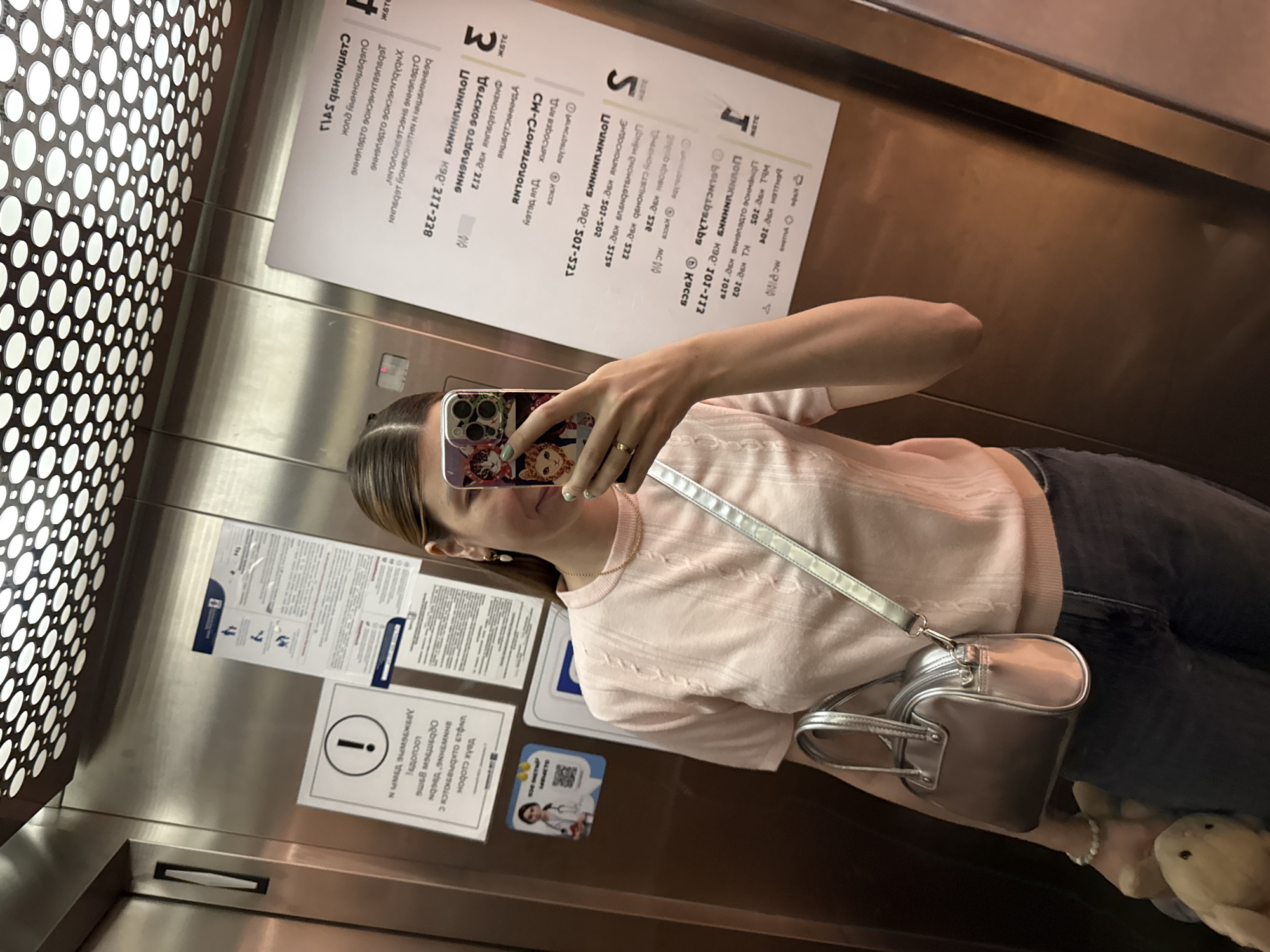 The image features a young woman taking a mirror selfie inside an elevator. She is partially visible, holding a smartphone with a decorative case, obscuring part of her face. She wears a light pink, short-sleeved top with a textured pattern and dark pants. A silver crossbody bag hangs from her shoulder, and a ring is visible on her finger. A light-colored stuffed animal is partially visible near her feet at the bottom right.
The elevator interior consists of reflective metal walls, possibly stainless steel, and a perforated panel on the left side. Numerous informational signs, predominantly in Russian, are attached to the walls. These signs appear to list different departments or services with corresponding room numbers, such as "211-228," "501-302," and "101-111." Visible Russian text includes "Детское отделение" (Children's Department) and "кровь" (blood), suggesting the setting is likely a medical facility or a building housing healthcare services. One sign displays an exclamation mark within a circle, indicating a warning or important information, and another features a QR code with an image resembling a doctor, potentially for service access or information. The scene is brightly lit, reflecting the internal lighting of the elevator.