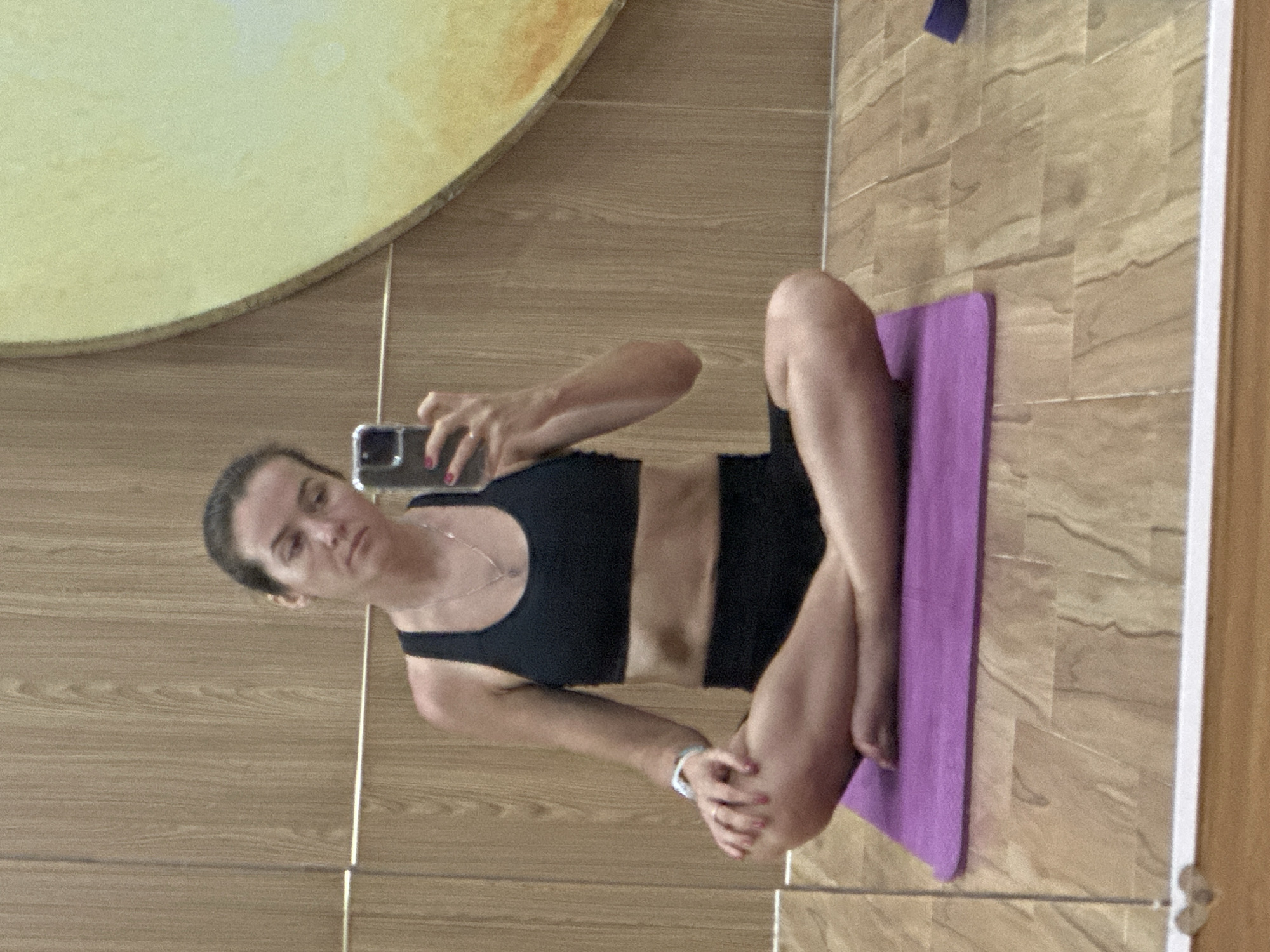 In Bac Nha Trang Ward, Vietnam, a woman in athletic wear sits cross-legged on a purple yoga mat in a room with wood-paneled walls. She is holding a smartphone in her right hand, taking a mirror selfie. The woman has her hair pulled back and is wearing a black sports bra and black shorts. A thin silver necklace is visible around her neck. A white fitness tracker is on her left wrist. The floor is made of wood planks, and a large, circular artwork with yellow and orange hues is partially visible on the wall to her right.