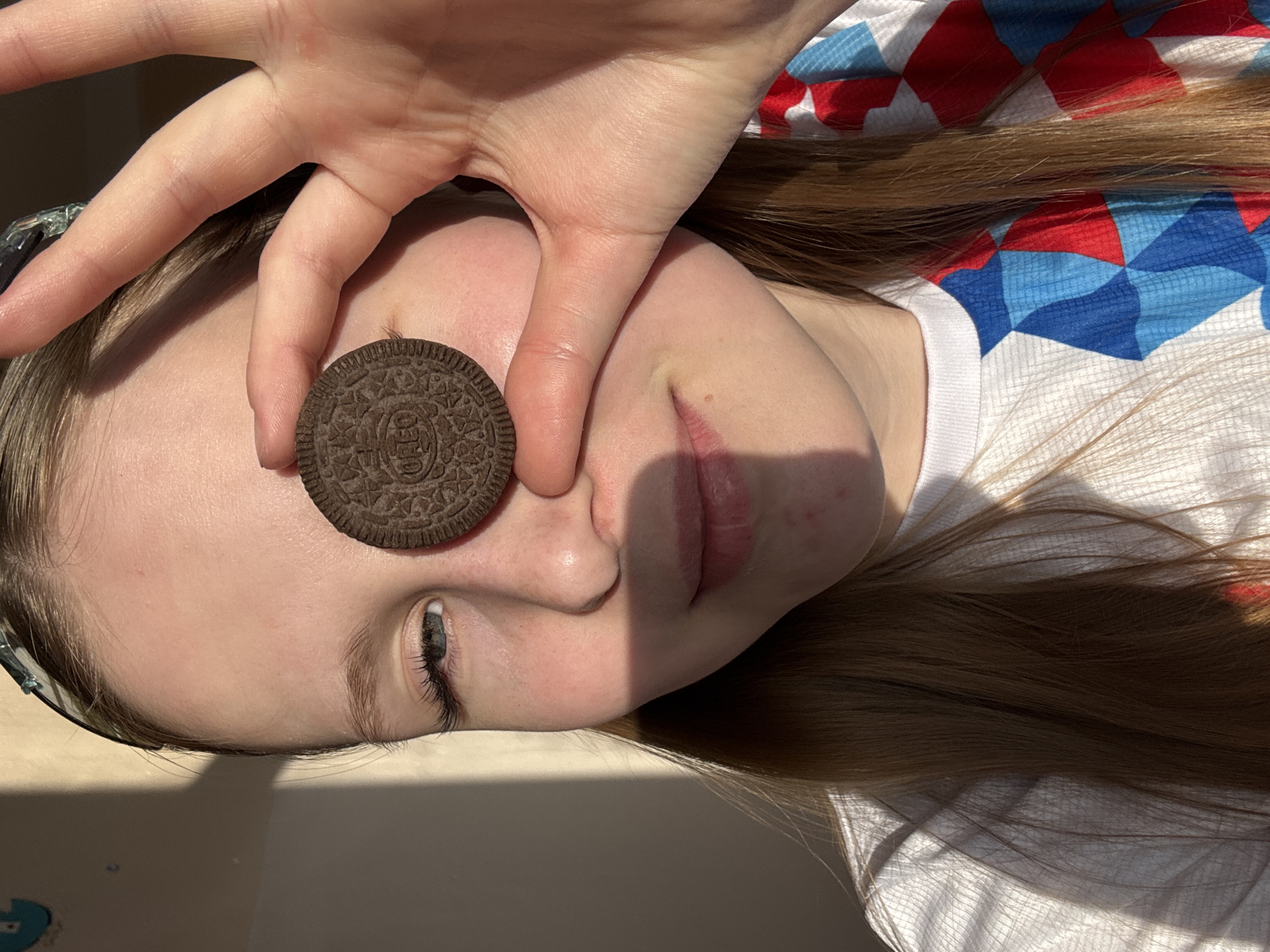 A close-up, horizontal, medium-shot depicts a fair-skinned individual with long, light brown hair lying down. The individual is wearing a white shirt with a red, white, and blue diamond pattern. A hand is holding a chocolate Oreo cookie in front of the individual's left eye. The location is not discernible as Yuzhnyy, Russia.