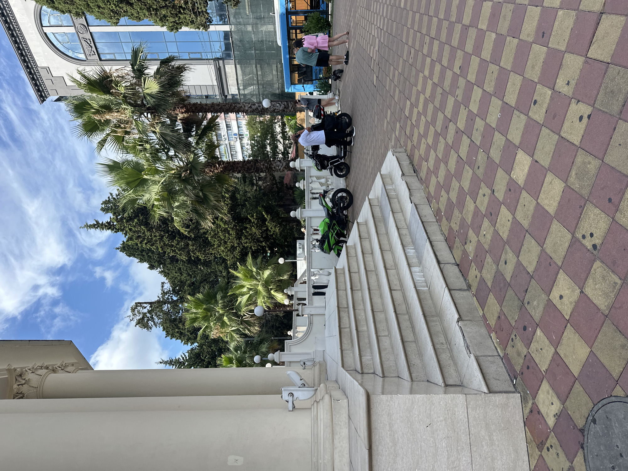 In front of a white neoclassical building with large columns and a wide, multi-tiered staircase, a checkered red and yellow tiled plaza is visible. Several palm trees and dark green conifers are scattered throughout the scene, with a large, modern building with a distinctive arched blue glass window in the background. On the plaza, two motorcycles, one bright green and the other black, are parked near the base of the stairs. Three individuals are present: a man in a white shirt and black pants stands by the black motorcycle, holding a phone to his ear. A woman in a grey sweatshirt and black shorts stands near him, looking at her phone. Further to the right, a man in a green t-shirt and black shorts stands next to a woman in a pink dress, both facing away from the camera. An electric scooter is positioned between them. A blue bus with large windows is partially visible on the far right of the frame. The sky above is blue with scattered white clouds. The scene appears to be in Sochi, Russia, given the architecture and vegetation typical of the region.